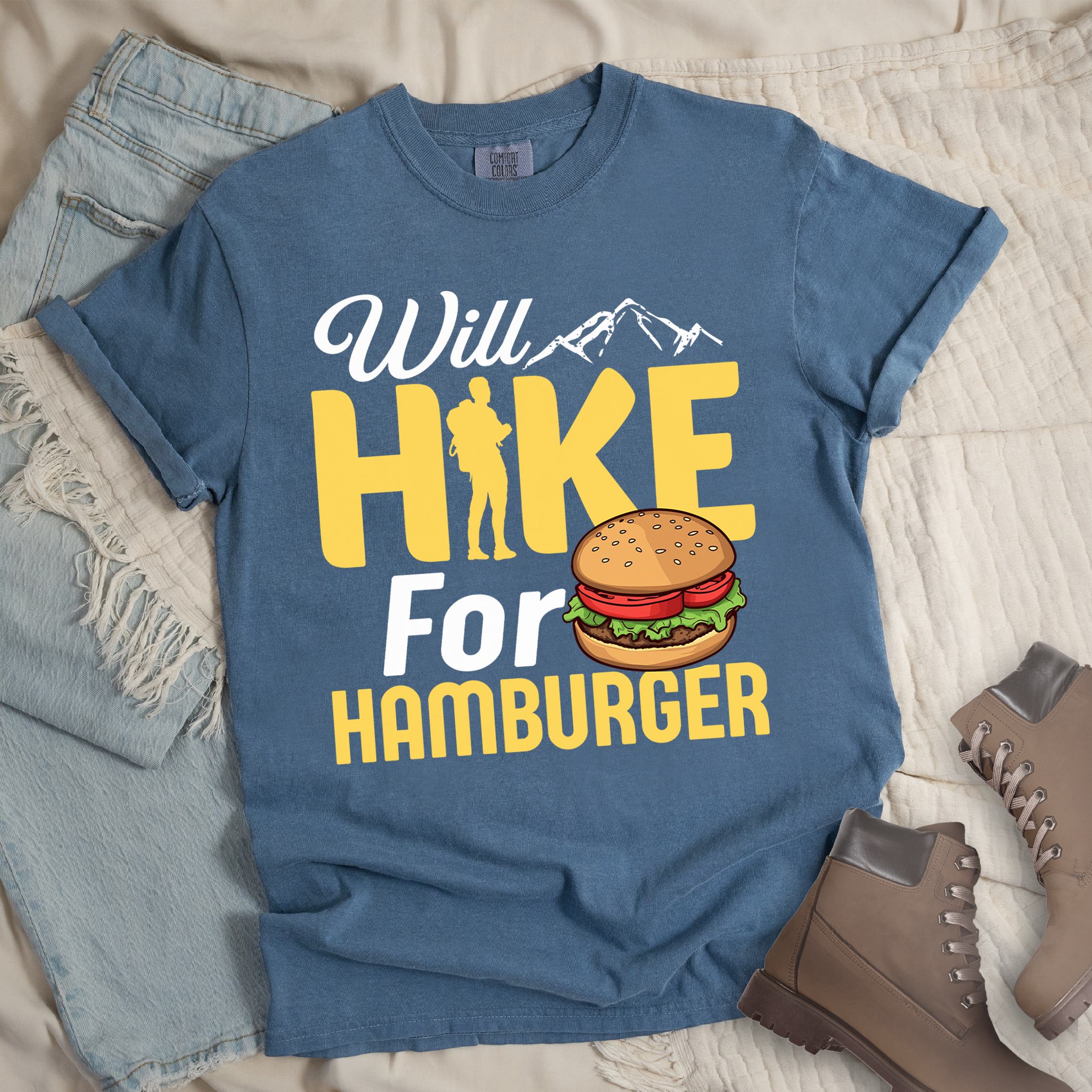 Dark blue t-shirt with text "Will HIKE For HAMBURGER". "Will" appears in white script with small mountain silhouettes. "HIKE" is in large yellow letters with a hiker silhouette replacing the letter "I". "For" text, and "HAMBURGER" at the bottom. Next to "For" and above "HAMBURGER" is a colorful illustration of a hamburger with sesame seed bun, lettuce, tomato, and patty.Features a classic crew neck and casual rolled sleeves, styled alongside light wash denim and brown hiking boots