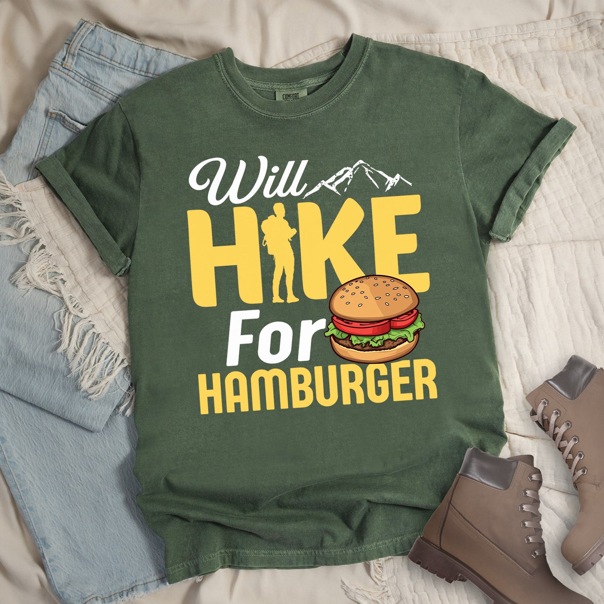 Dark green t-shirt with text "Will HIKE For HAMBURGER". "Will" appears in white script with small mountain silhouettes. "HIKE" is in large yellow letters with a hiker silhouette replacing the letter "I". "For" text, and "HAMBURGER" at the bottom. Next to "For" and above "HAMBURGER" is a colorful illustration of a hamburger with sesame seed bun, lettuce, tomato, and patty.Features a classic crew neck and casual rolled sleeves, styled alongside light wash denim and brown hiking boots