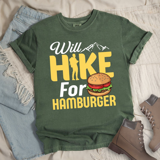 Dark green t-shirt with text "Will HIKE For HAMBURGER". "Will" appears in white script with small mountain silhouettes. "HIKE" is in large yellow letters with a hiker silhouette replacing the letter "I". "For" text, and "HAMBURGER" at the bottom. Next to "For" and above "HAMBURGER" is a colorful illustration of a hamburger with sesame seed bun, lettuce, tomato, and patty.Features a classic crew neck and casual rolled sleeves, styled alongside light wash denim and brown hiking boots