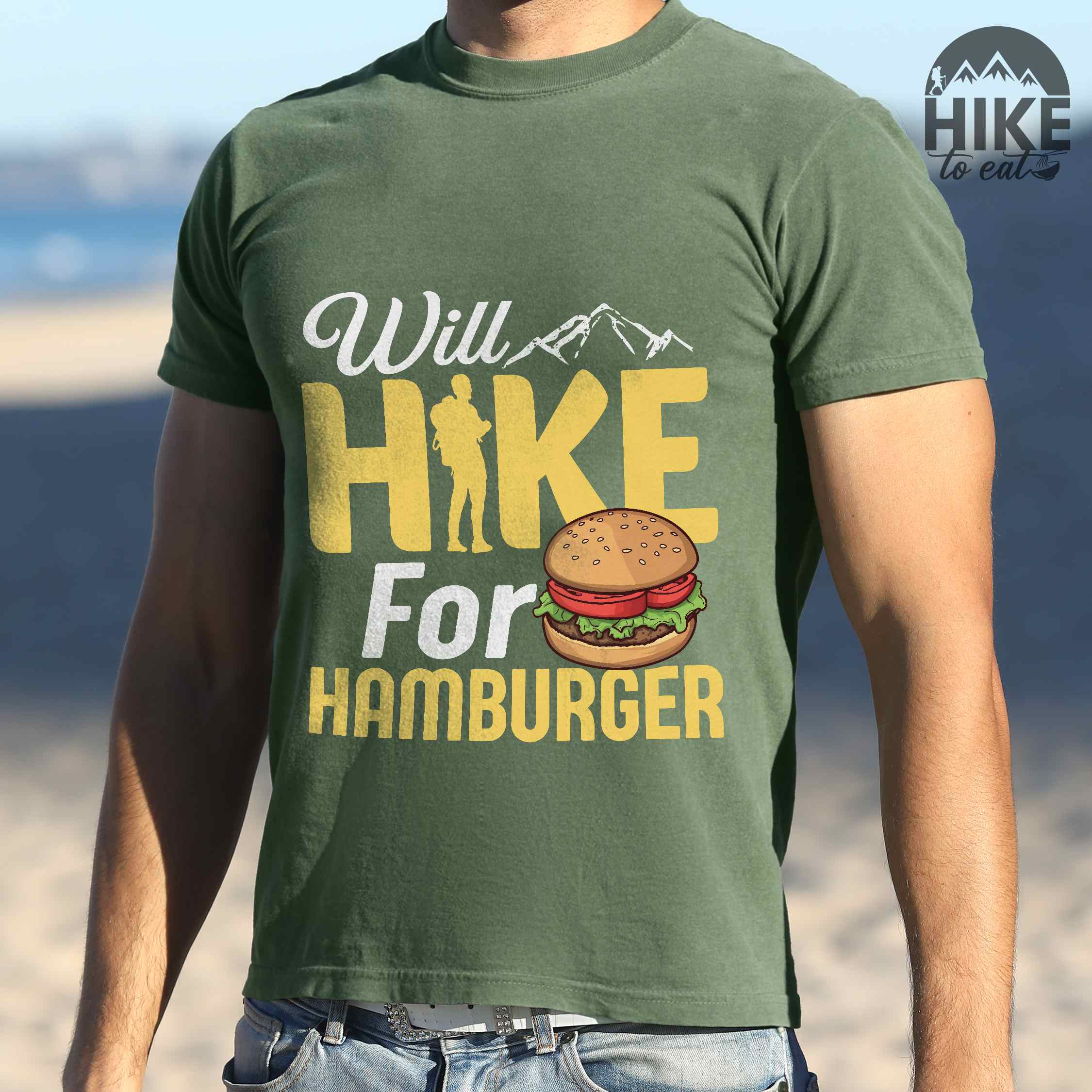 Man wearing moss "Will Hike for Hamburger" colored vintage tee with bold typography, birds, mountains, illustrated juicy hamburger, and backpacker for burger enthusiasts.