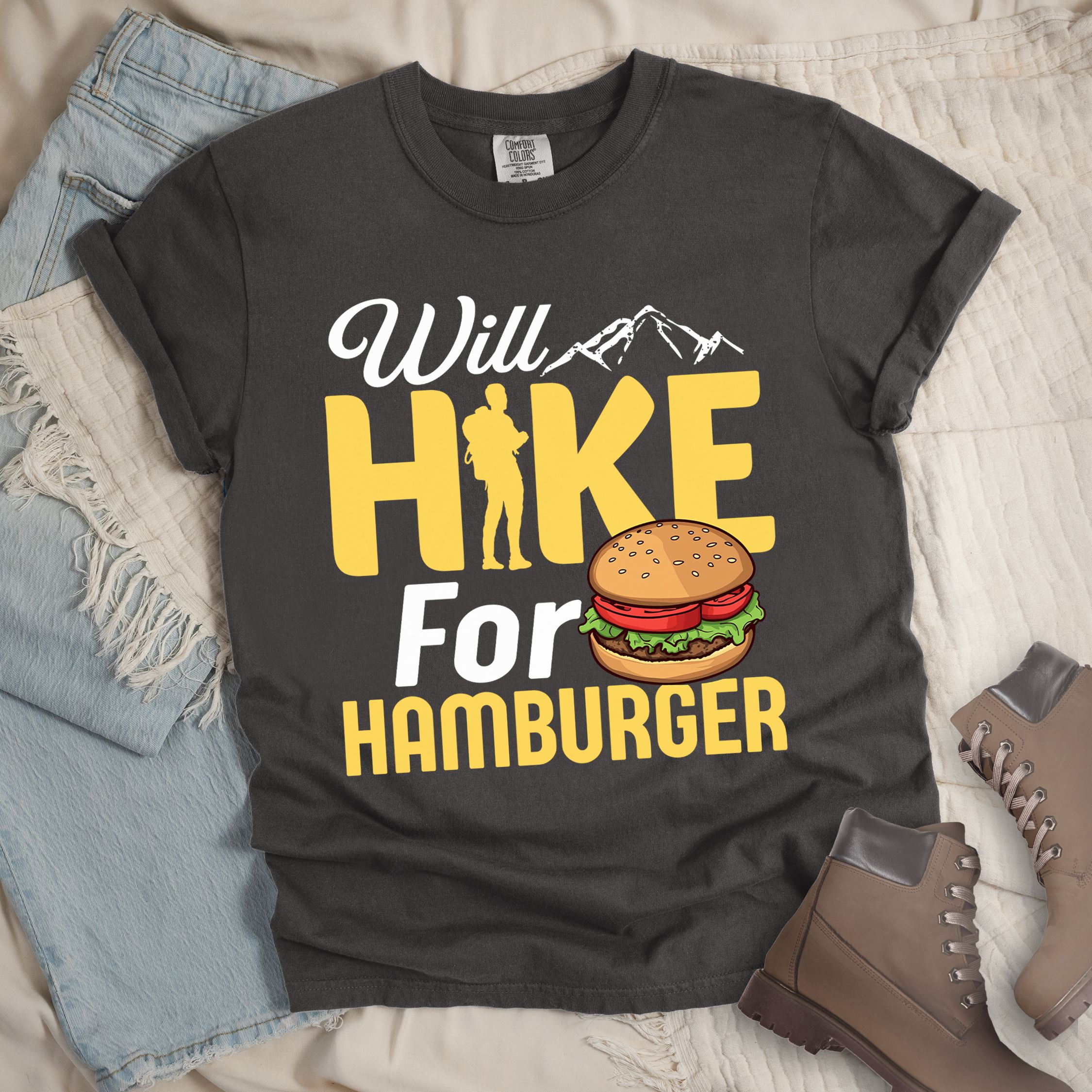 Dark grey t-shirt with text "Will HIKE For HAMBURGER". "Will" appears in white script with small mountain silhouettes. "HIKE" is in large yellow letters with a hiker silhouette replacing the letter "I". "For" text, and "HAMBURGER" at the bottom. Next to "For" and above "HAMBURGER" is a colorful illustration of a hamburger with sesame seed bun, lettuce, tomato, and patty.Features a classic crew neck and casual rolled sleeves, styled alongside light wash denim and brown hiking boots