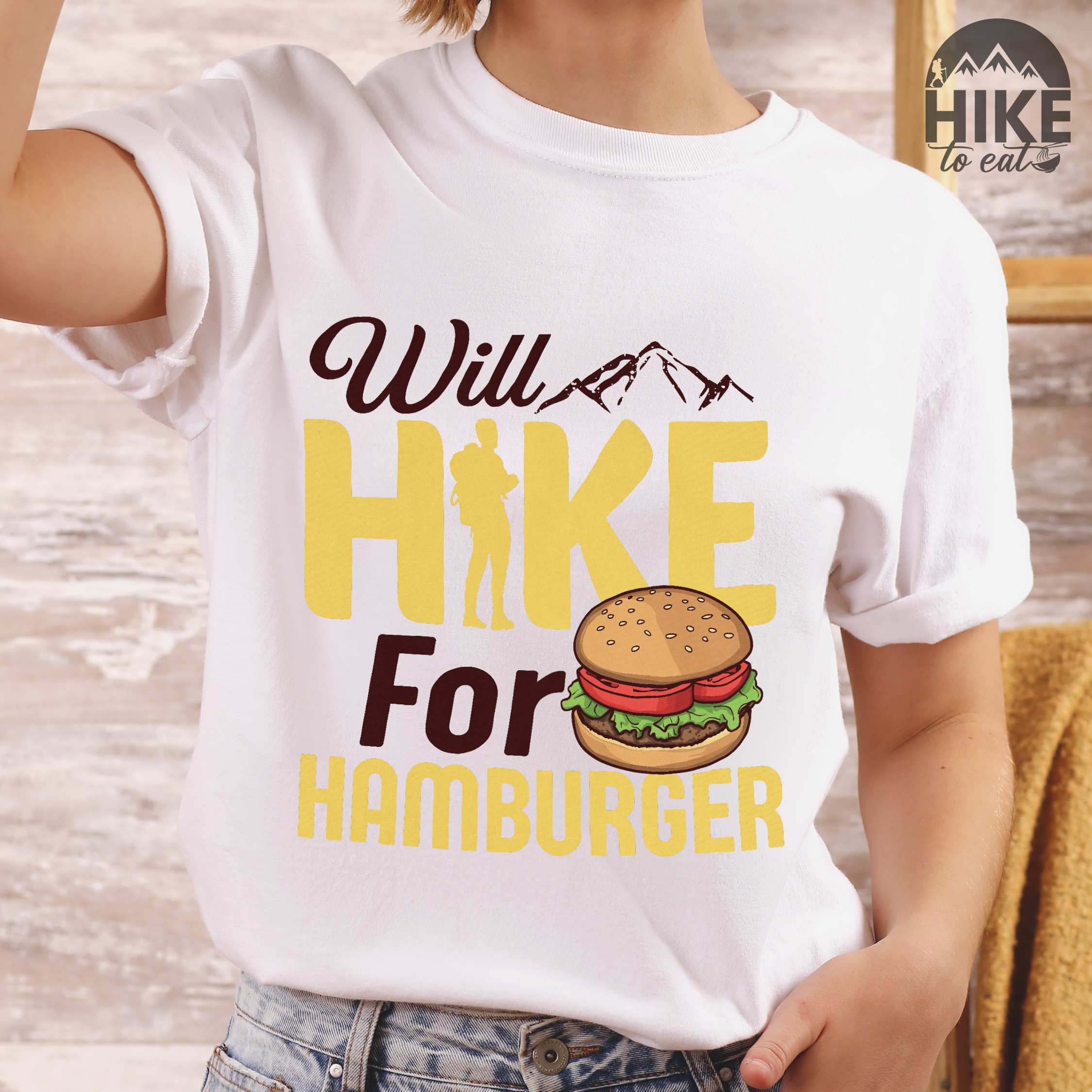 Woman wearing white "Will Hike for Hamburger" colored vintage tee with bold typography, birds, mountains, illustrated juicy hamburger, and backpacker for burger enthusiasts.