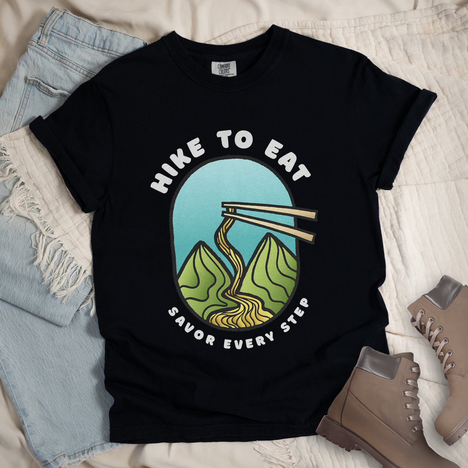 A black t-shirt with white text "HIKE TO EAT" arched at the top and "SAVOR EVERY STEP" at the bottom. The center features an oval graphic with a turquoise blue sky background, green mountain peaks, and chopsticks lifting ramen noodles that flow like a trail between the mountains. The shirt features a classic crew neck and casual rolled sleeves, styled alongside light wash denim and brown hiking boots.