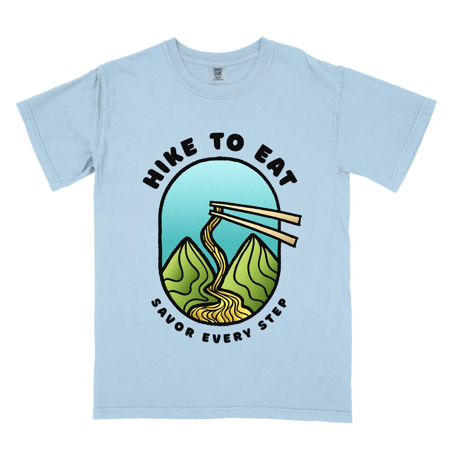 A chambray (light blue) t-shirt with black text "HIKE TO EAT" arched at the top and "SAVOR EVERY STEP" at the bottom. The center features an oval graphic with a turquoise blue sky background, green mountain peaks, and chopsticks lifting ramen noodles that flow like a trail between the mountains. 