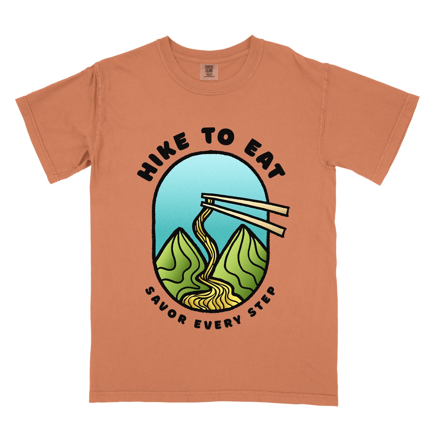 A yam (brown) t-shirt with black text "HIKE TO EAT" arched at the top and "SAVOR EVERY STEP" at the bottom. The center features an oval graphic with a turquoise blue sky background, green mountain peaks, and chopsticks lifting ramen noodles that flow like a trail between the mountains. 