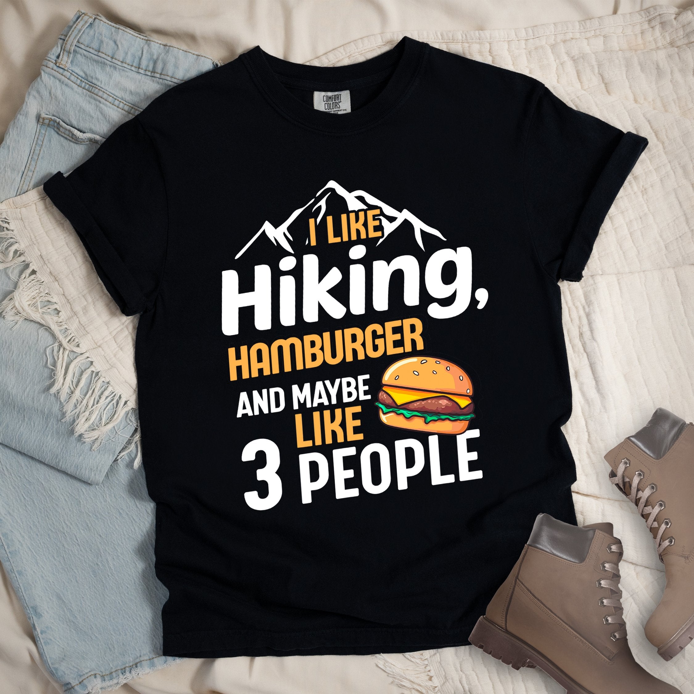 Black t-shirt with text "I LIKE Hiking, HAMBURGER AND MAYBE LIKE 3 PEOPLE". mountain silhouettes appear at the top. An illustration of a hamburger with sesame seed bun, cheese, lettuce and patty appears next to the word "LIKE". The design combines hiking and introvert humor.
