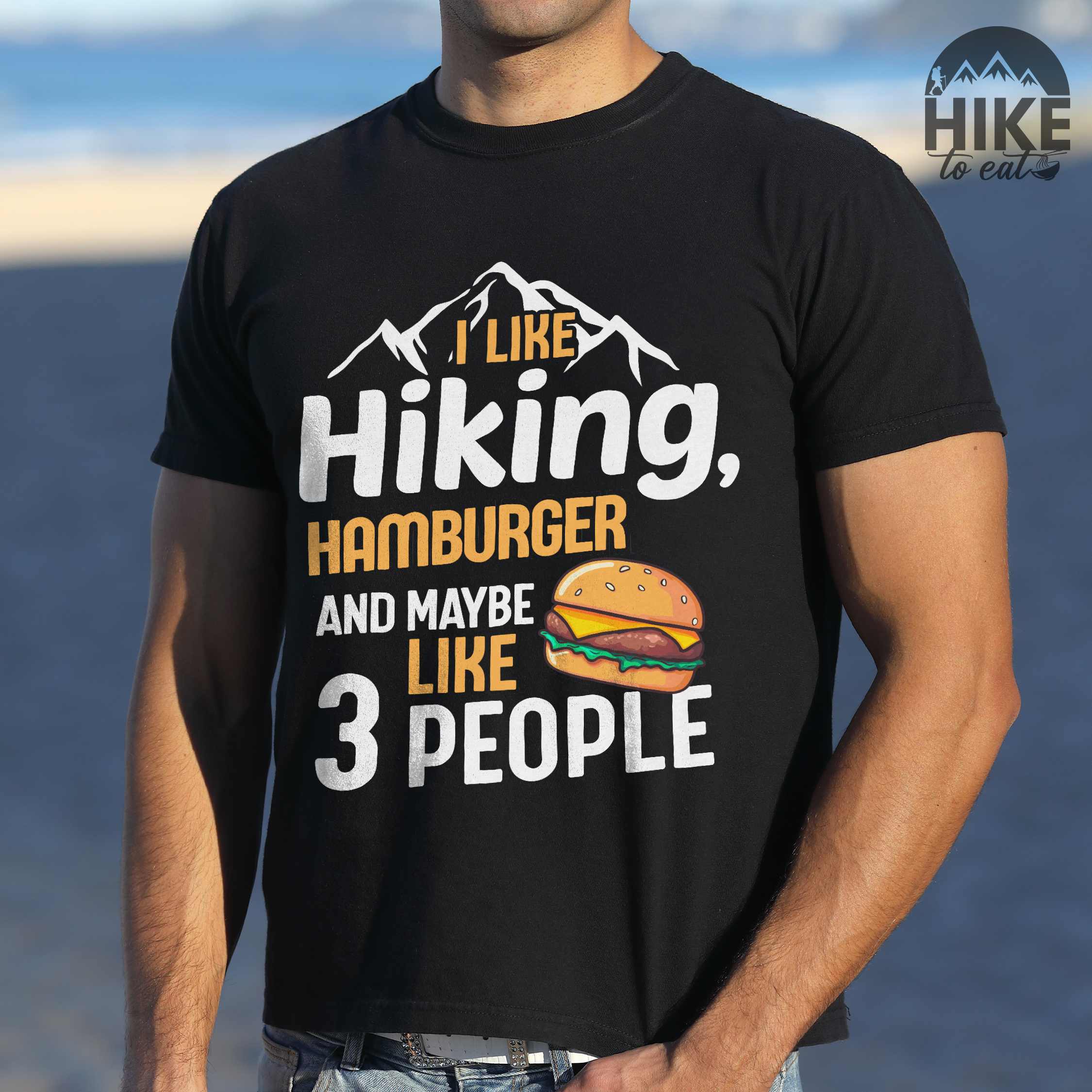 Man wearing black "I like hiking, hamburger and maybe like three people" colored vintage tee with bold typography, birds, mountains, illustrated juicy hamburger, and backpacker for burger enthusiasts.