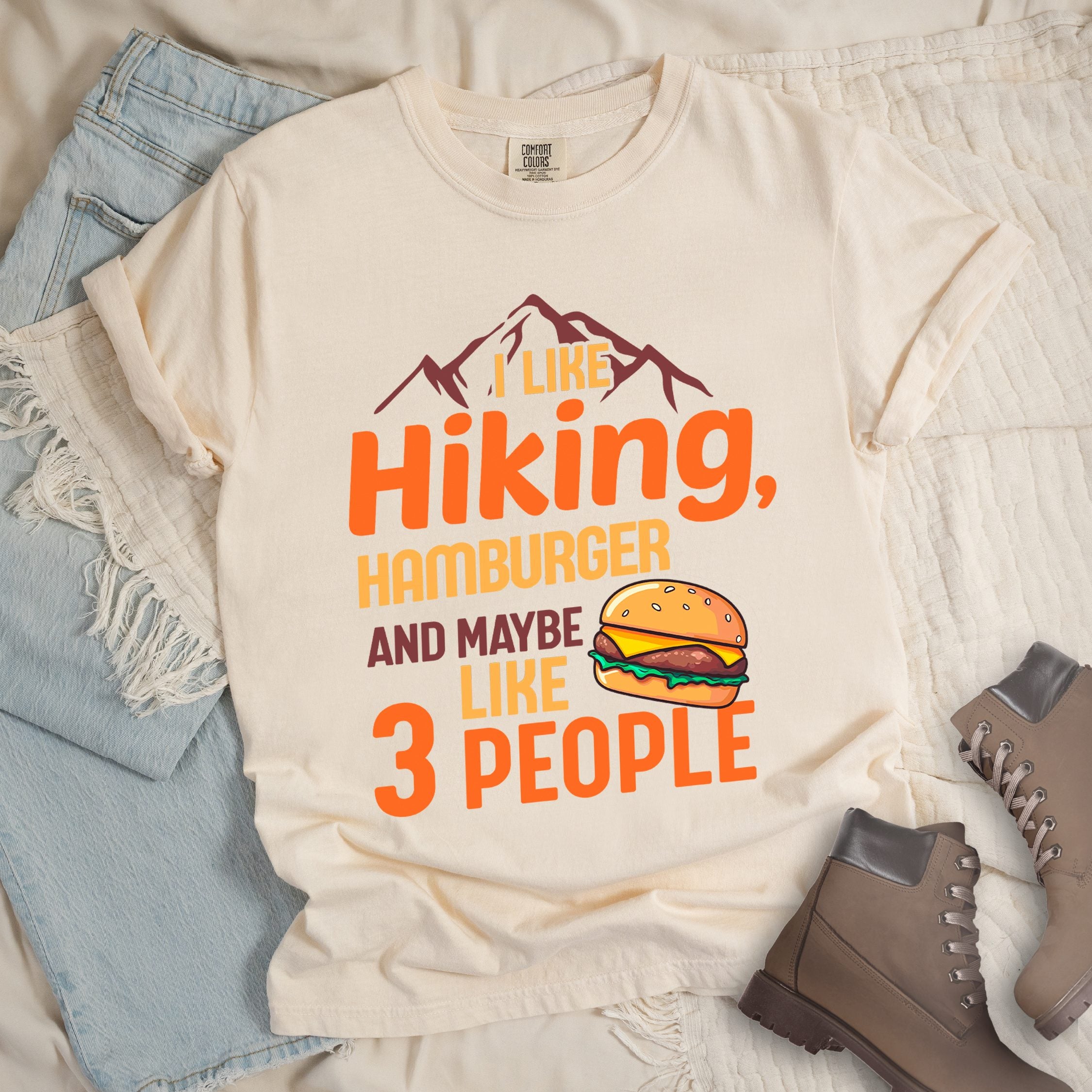 Ivory t-shirt with text "I LIKE Hiking, HAMBURGER AND MAYBE LIKE 3 PEOPLE". mountain silhouettes appear at the top. An illustration of a hamburger with sesame seed bun, cheese, lettuce and patty appears next to the word "LIKE". The design combines hiking and introvert humor.