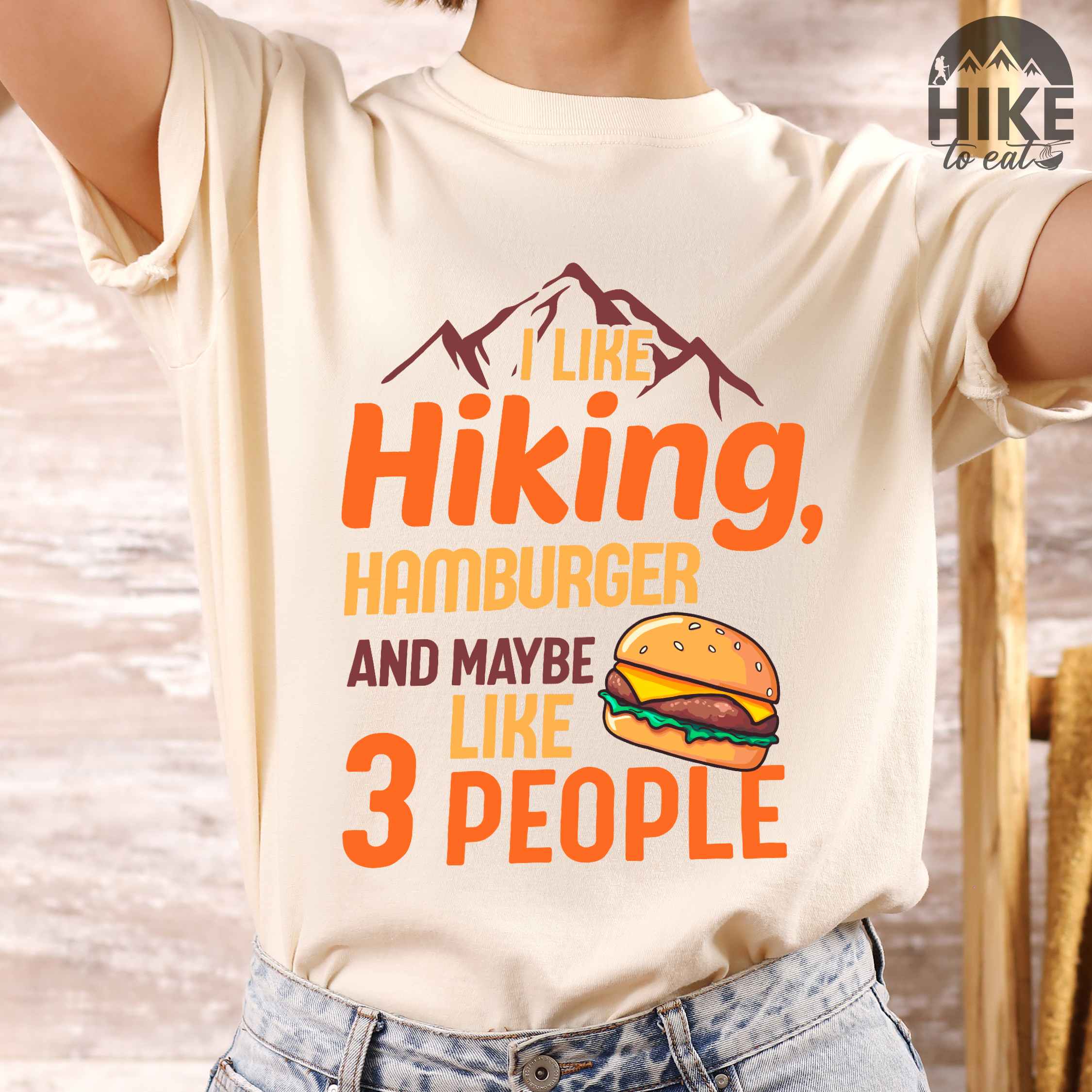 Woman wearing ivory "I like hiking, hamburger and maybe like three people" colored vintage tee with bold typography, birds, mountains, illustrated juicy hamburger, and backpacker for burger enthusiasts.