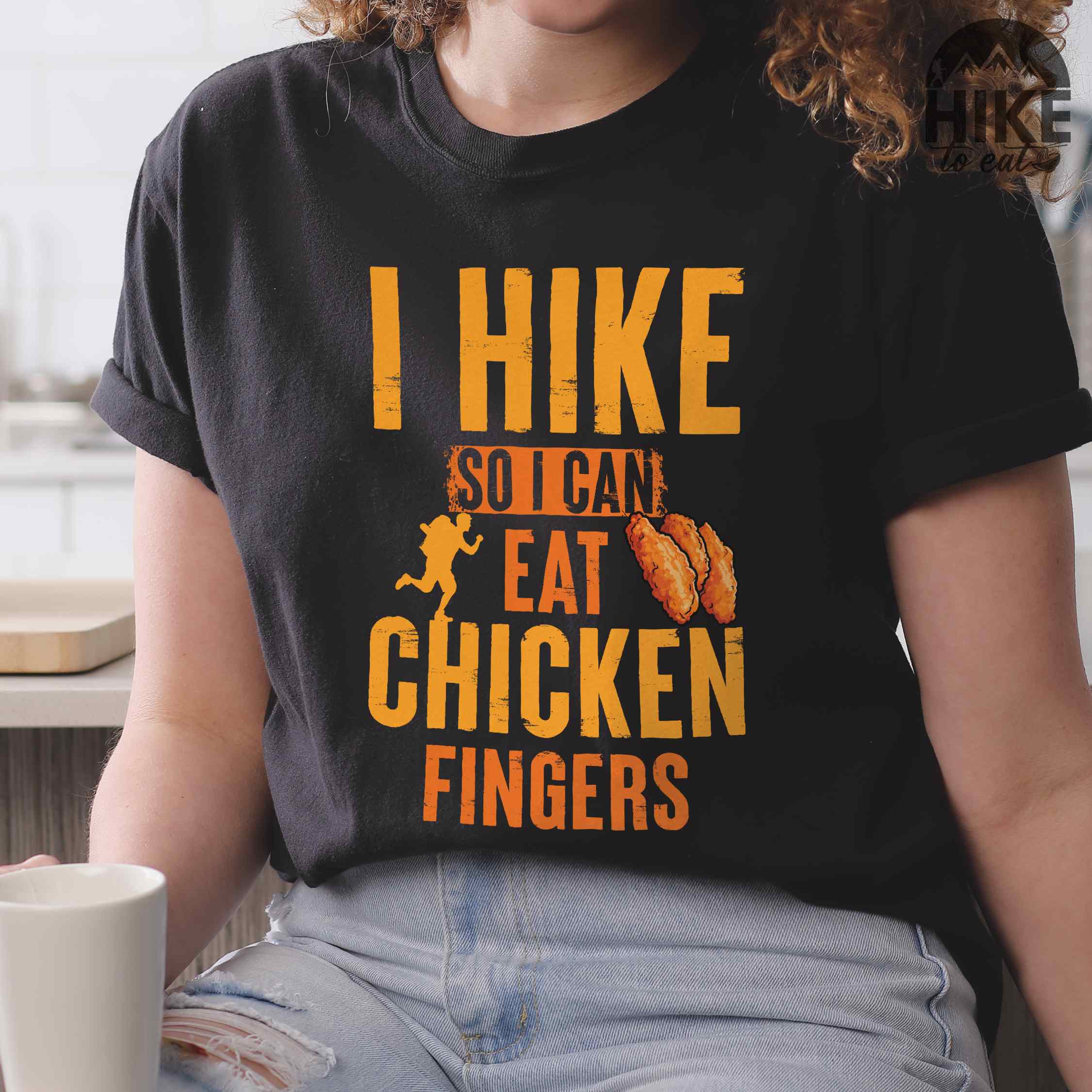 Woman wearing black "I Hike so I can eat Chicken Fingers" colored vintage tee with bold typography, illustrated crispy fried chicken fingers, perfect for comfort food lovers.