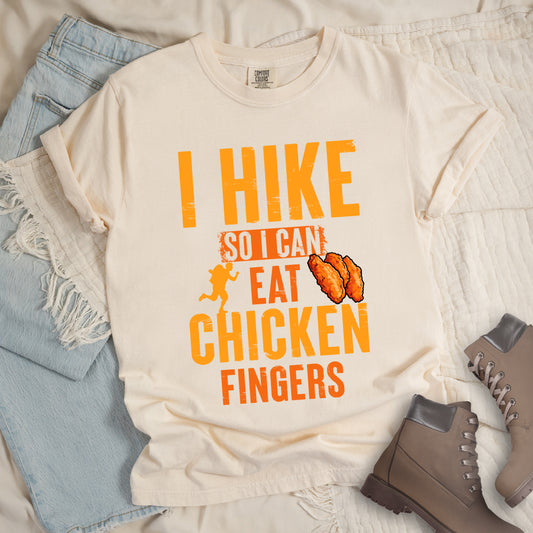 Ivory (Cream White) t-shirt with orange distressed text reading "I HIKE SO I CAN EAT CHICKEN FINGERS". All text appears in gradient orange tones from yellow-orange to deeper orange. A small hiker silhouette appears after "SO I CAN", and an illustration of three breaded chicken fingers/tenders appears next to "EAT". The design presents hiking as justification for enjoying comfort food, with a playful, casual aesthetic.