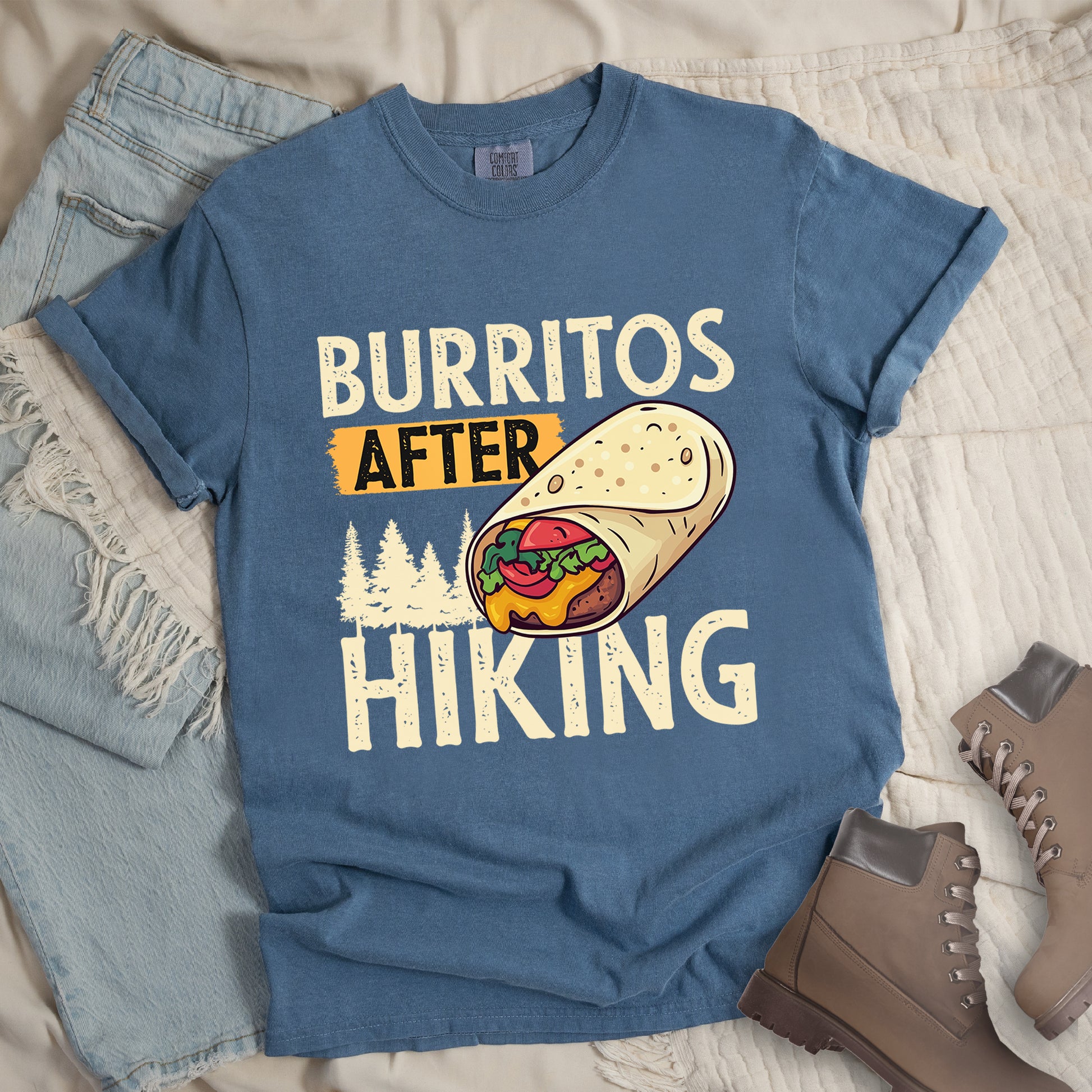 Blue Jean (Denim Blue) t-shirt with distressed text reading "BURRITOS AFTER HIKING". "BURRITOS" appears at the top in cream/off-white letters, "AFTER" in black text on an orange background in the middle, and "HIKING" in matching cream letters at the bottom. Between the text is an illustration of a burrito with visible fillings (beans, cheese, lettuce, tomato) and pine tree silhouettes in cream color. The design has a vintage, weathered appearance.