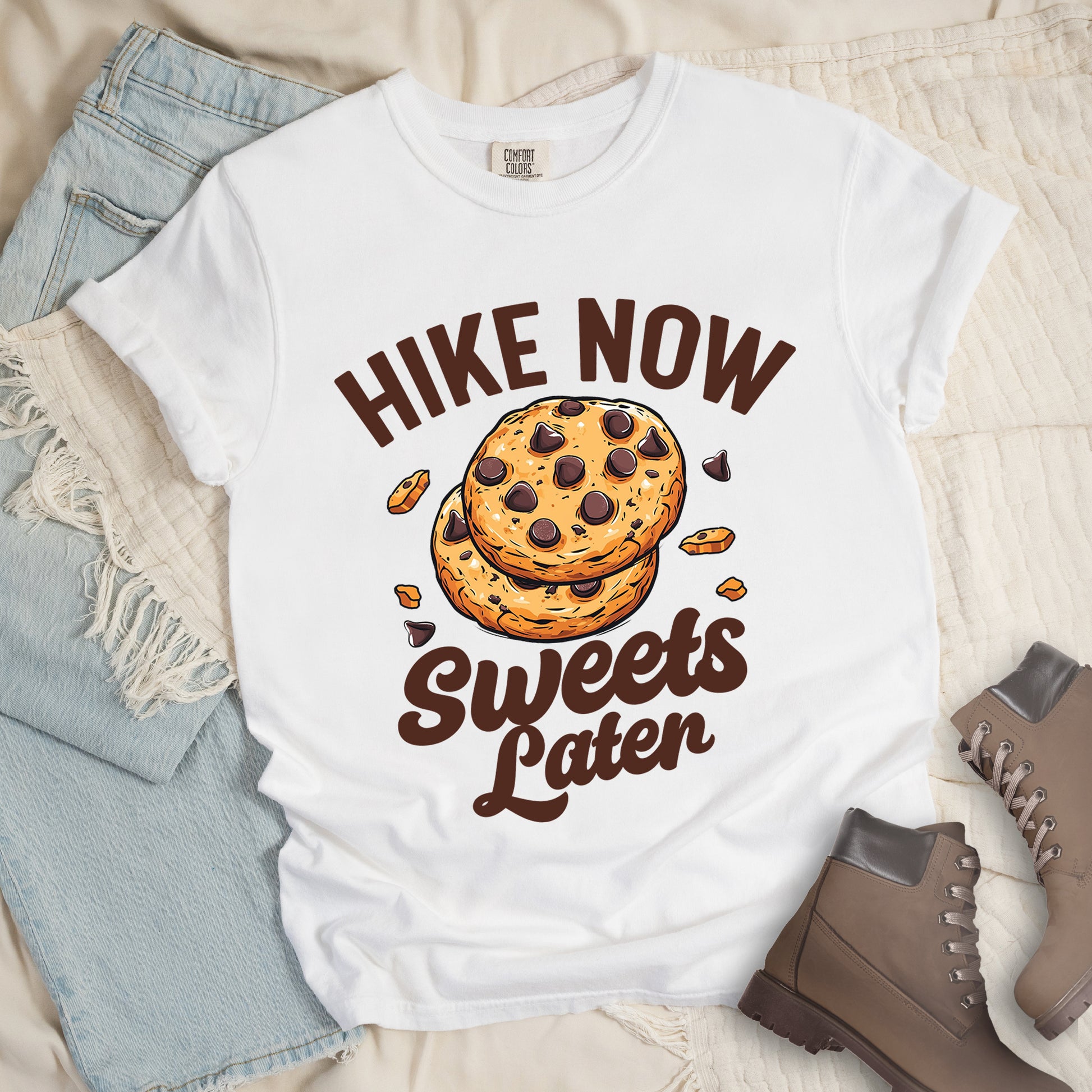 White t-shirt with "HIKE NOW Sweets Later" text design. "HIKE NOW" appears in brown capital letters arched at the top, and "Sweets Later" in brown script at the bottom. In the center is an illustration of two chocolate chip cookies stacked on top of each other with some cookie crumbs and chocolate chunks scattered around them. The design emphasizes hiking first with dessert as a reward afterward.
