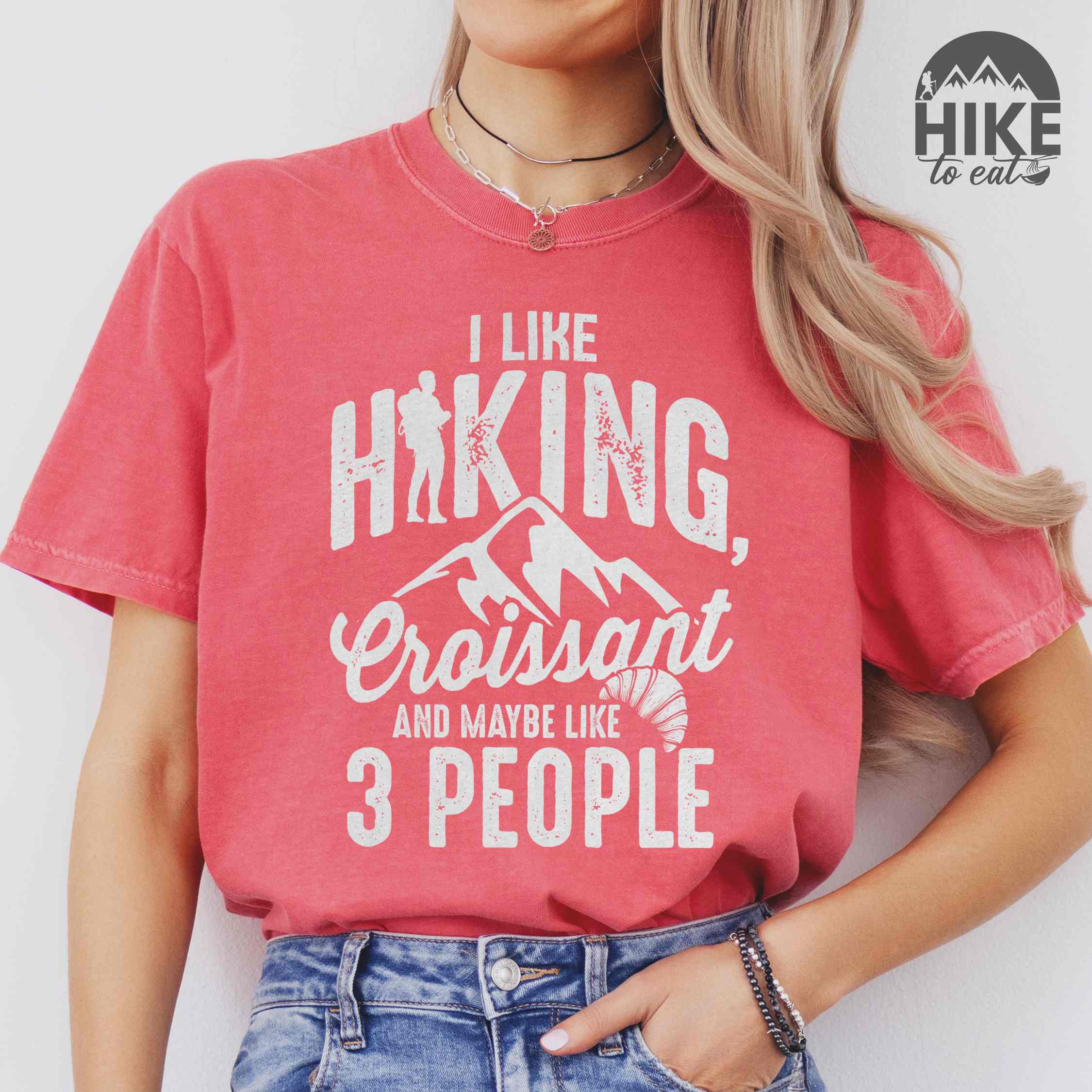Woman wearing watermelon "I Like hiking, croissant and maybe like 3 people" colored vintage tee with bold typography, birds, mountains, illustrated croissant, and backpacker for pastry enthusiasts.
