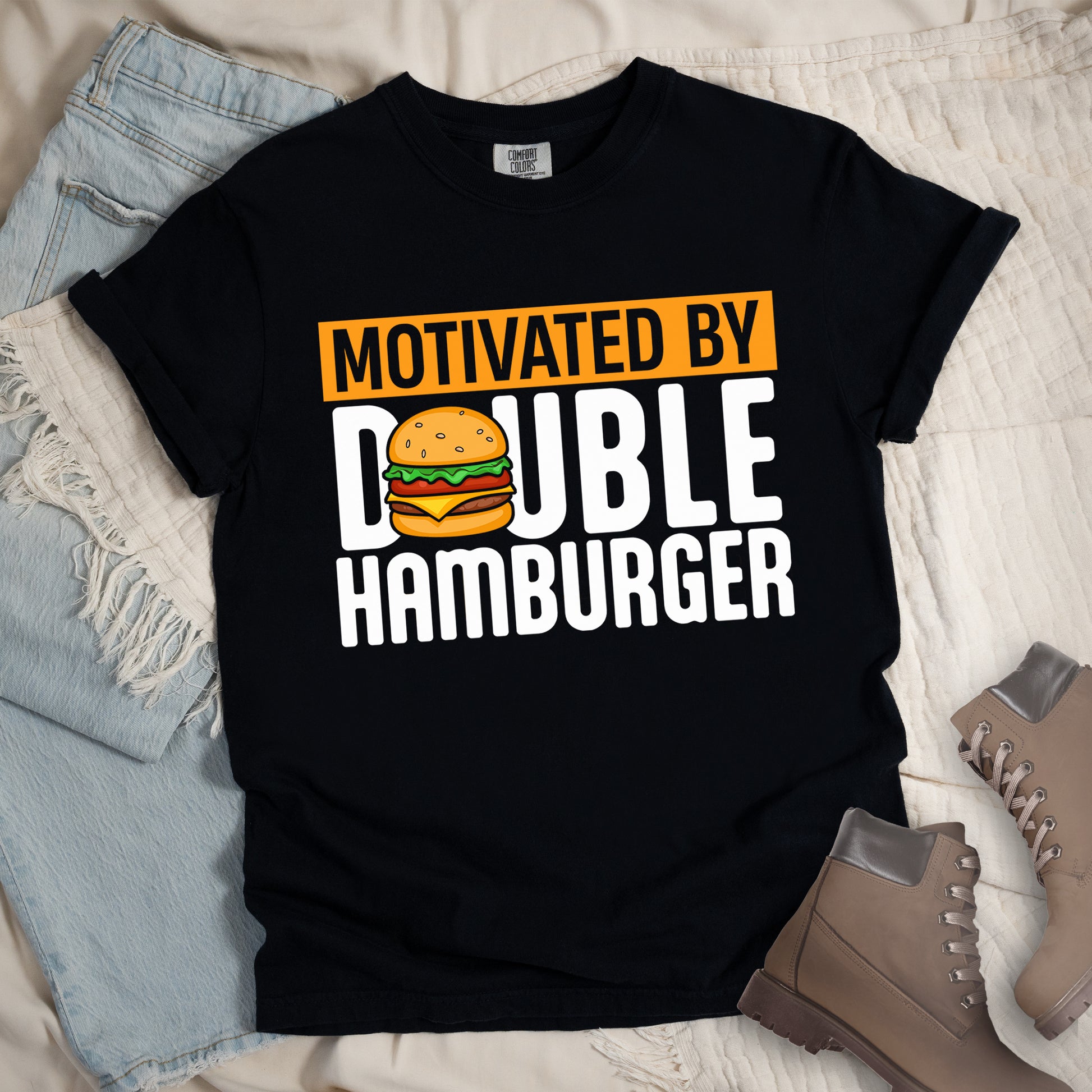 Black t-shirt with text "MOTIVATED BY DOUBLE HAMBURGER". "MOTIVATED BY" appears in black text on an orange rectangular background at the top. "DOUBLE HAMBURGER" appears in large white letters below, with a colorful hamburger illustration replacing the letter "O" in "DOUBLE". The hamburger graphic shows a sesame seed bun, lettuce, tomato, cheese, and beef patty.