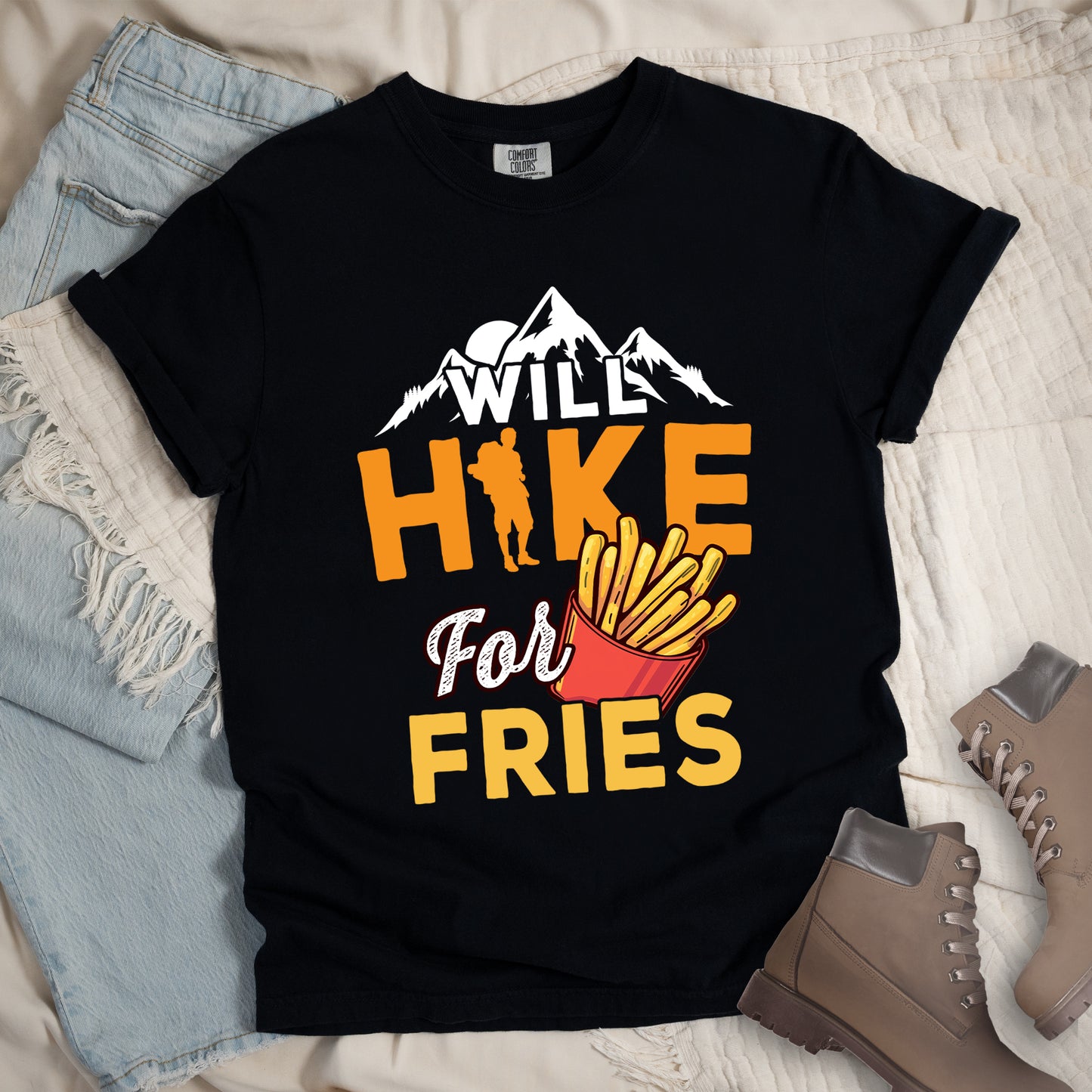 Black t-shirt with text "WILL HIKE For FRIES". White mountain silhouettes appear above "WILL" in white letters. "HIKE" is in large orange letters with a hiker silhouette replacing the letter "I". "For" appears in white script, and "FRIES" in yellow at the bottom. Next to "For" and above "FRIES" is an illustration of french fries in a red container. The design combines hiking motivation with food rewards.