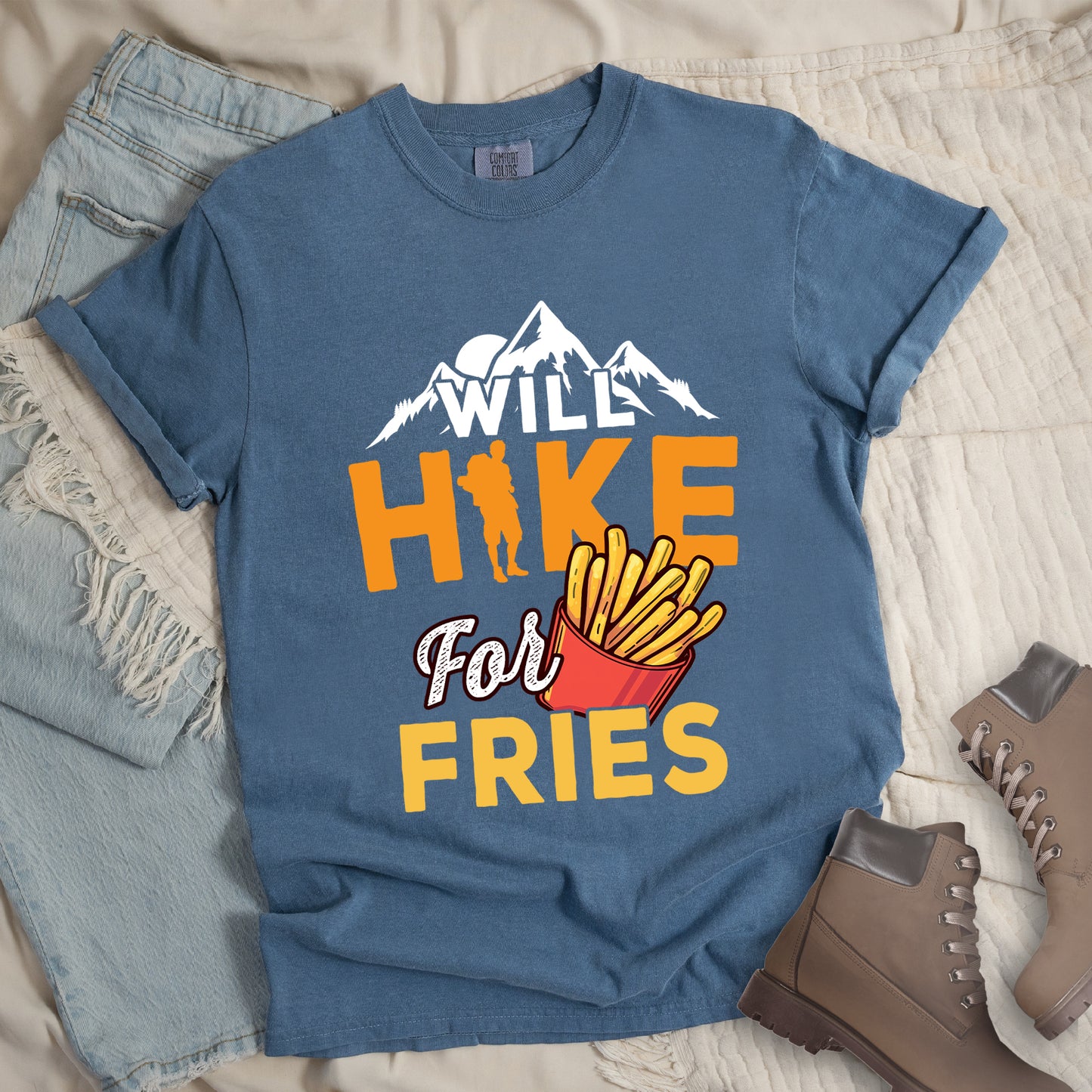 Blue Jean (Denim Blue) t-shirt with text "WILL HIKE For FRIES". White mountain silhouettes appear above "WILL" in white letters. "HIKE" is in large orange letters with a hiker silhouette replacing the letter "I". "For" appears in white script, and "FRIES" in yellow at the bottom. Next to "For" and above "FRIES" is an illustration of french fries in a red container. The design combines hiking motivation with food rewards.