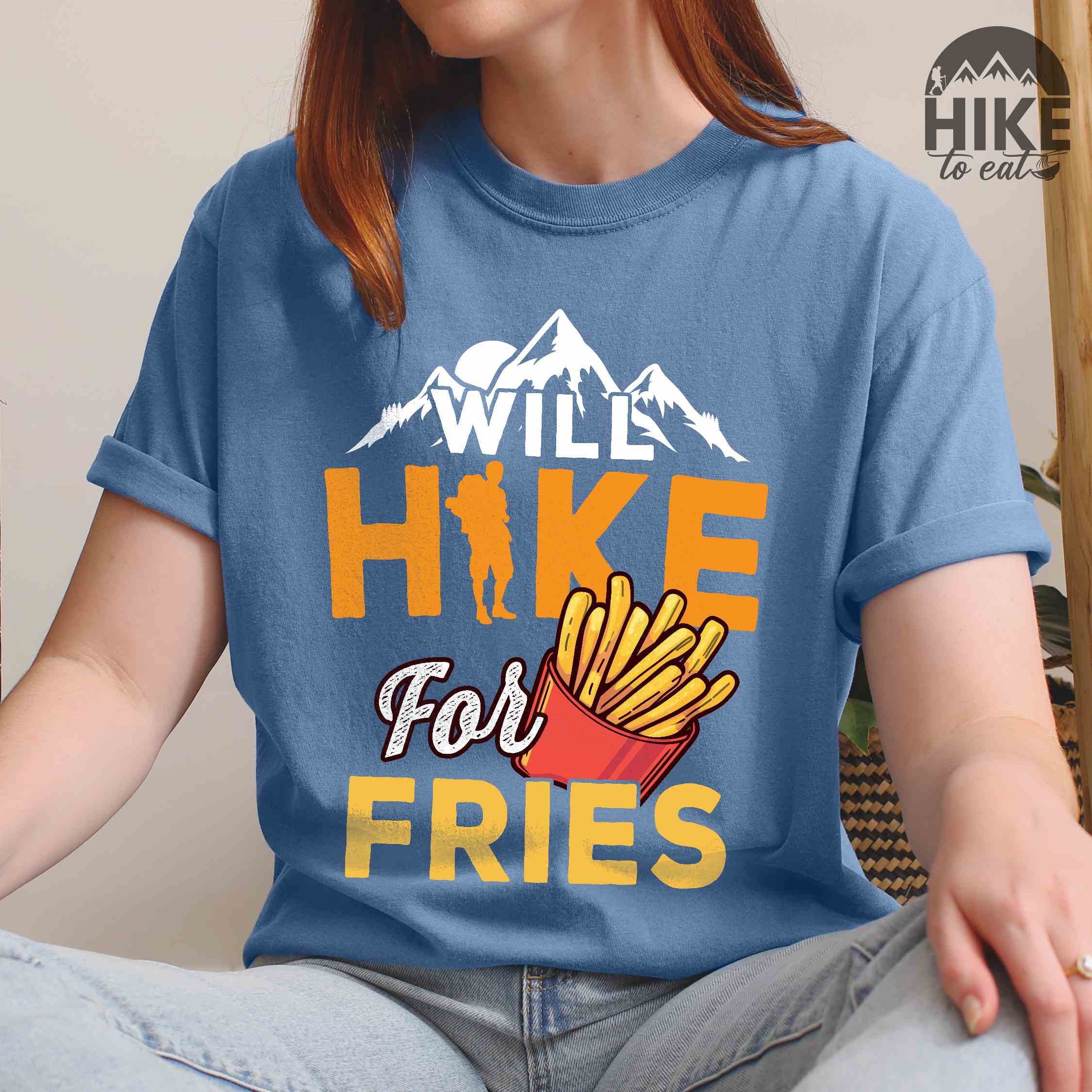 Woman wearing blue jean "Will Hike for Fries" colored vintage tee with bold typography, mountains, illustrated fries, and backpacker for comfort food enthusiasts.
