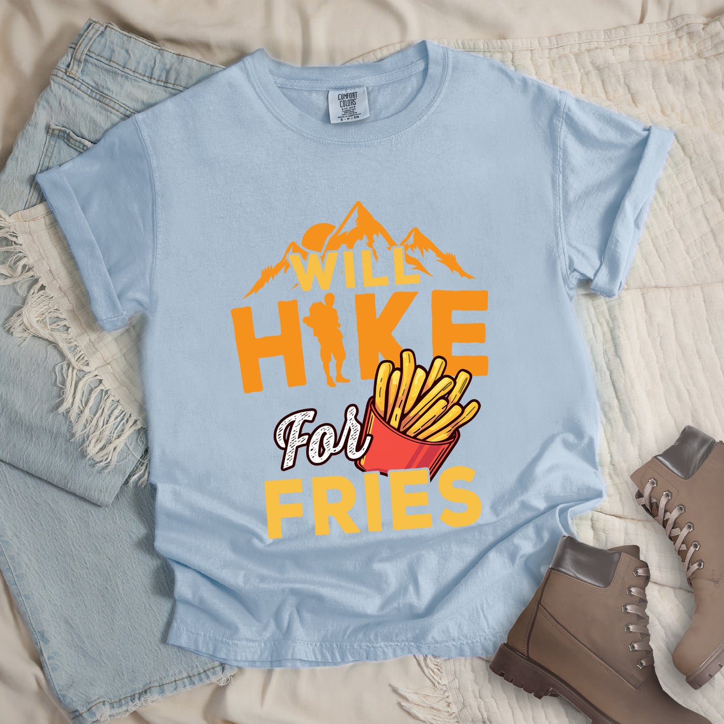 Chambray (Light Blue) t-shirt with text "WILL HIKE For FRIES". Orange mountain silhouettes appear above "WILL" in yellow letters. "HIKE" is in large orange letters with a hiker silhouette replacing the letter "I". "For" appears in white script, and "FRIES" in yellow at the bottom. Next to "For" and above "FRIES" is an illustration of french fries in a red container. The design combines hiking motivation with food rewards.