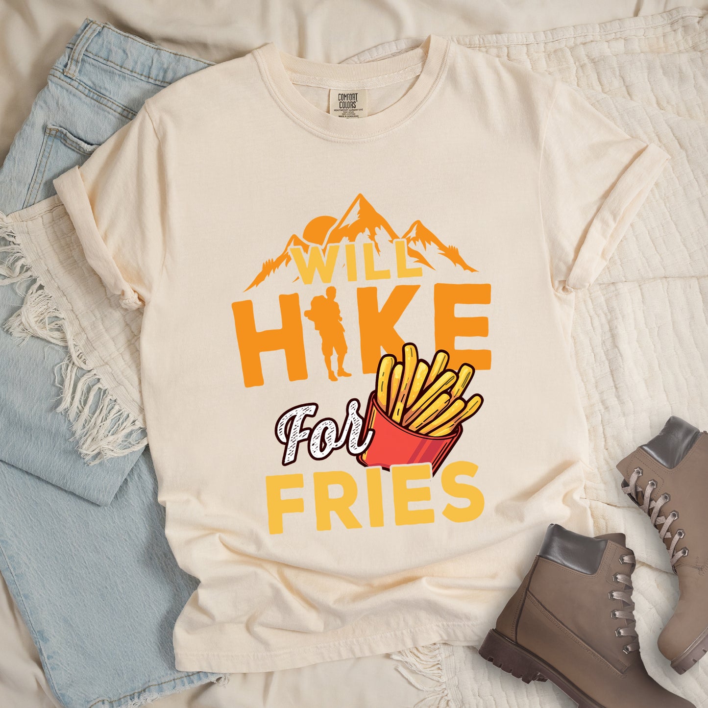 Ivory (Cream White) t-shirt with text "WILL HIKE For FRIES". Orange mountain silhouettes appear above "WILL" in yellow letters. "HIKE" is in large orange letters with a hiker silhouette replacing the letter "I". "For" appears in white script, and "FRIES" in yellow at the bottom. Next to "For" and above "FRIES" is an illustration of french fries in a red container. The design combines hiking motivation with food rewards.