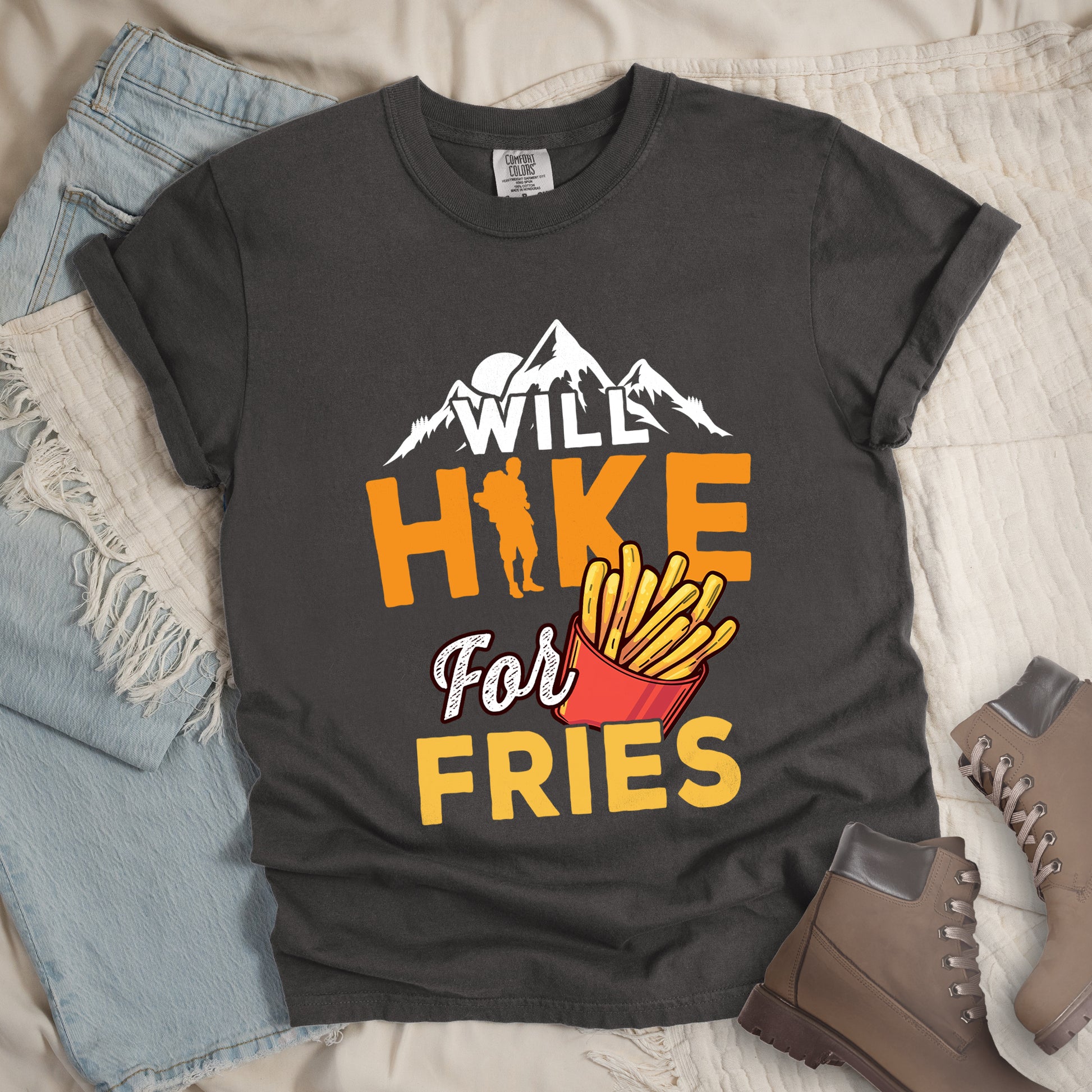 Pepper (Dark Gray) t-shirt with text "WILL HIKE For FRIES". White mountain silhouettes appear above "WILL" in white letters. "HIKE" is in large orange letters with a hiker silhouette replacing the letter "I". "For" appears in white script, and "FRIES" in yellow at the bottom. Next to "For" and above "FRIES" is an illustration of french fries in a red container. The design combines hiking motivation with food rewards.
