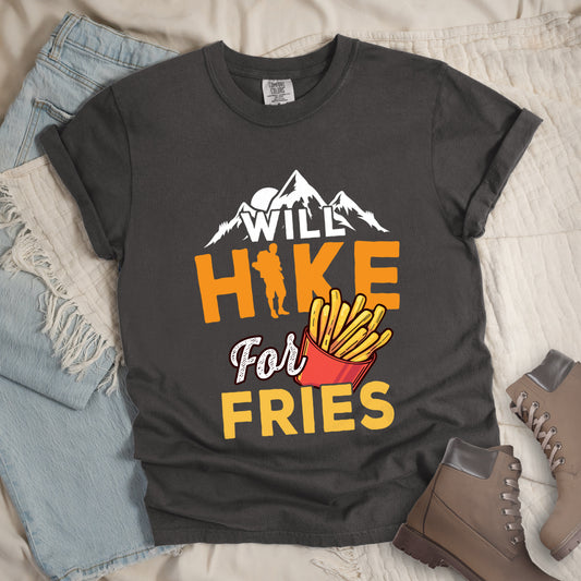 Pepper (Dark Gray) t-shirt with text "WILL HIKE For FRIES". White mountain silhouettes appear above "WILL" in white letters. "HIKE" is in large orange letters with a hiker silhouette replacing the letter "I". "For" appears in white script, and "FRIES" in yellow at the bottom. Next to "For" and above "FRIES" is an illustration of french fries in a red container. The design combines hiking motivation with food rewards.