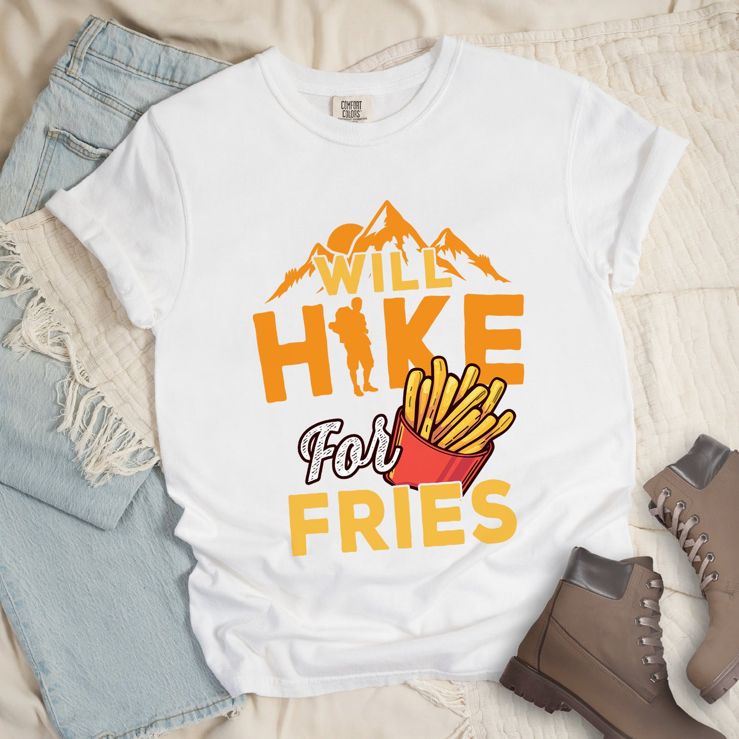 White t-shirt with text "WILL HIKE For FRIES". Orange mountain silhouettes appear above "WILL" in yellow letters. "HIKE" is in large orange letters with a hiker silhouette replacing the letter "I". "For" appears in white script, and "FRIES" in yellow at the bottom. Next to "For" and above "FRIES" is an illustration of french fries in a red container. The design combines hiking motivation with food rewards.