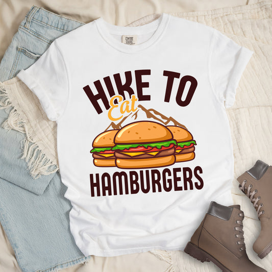 White t-shirt with text "HIKE TO Eat HAMBURGERS". "HIKE TO" appears in deep brown arched letters at the top, "Eat" in orange script in the middle, and "HAMBURGERS" in deep brown at the bottom. The center features an illustration of three hamburgers with buns, lettuce, tomato, cheese and beef patties, with brown mountain silhouettes behind them. The shirt features a classic crew neck and casual rolled sleeves, styled alongside light wash denim and brown hiking boots.