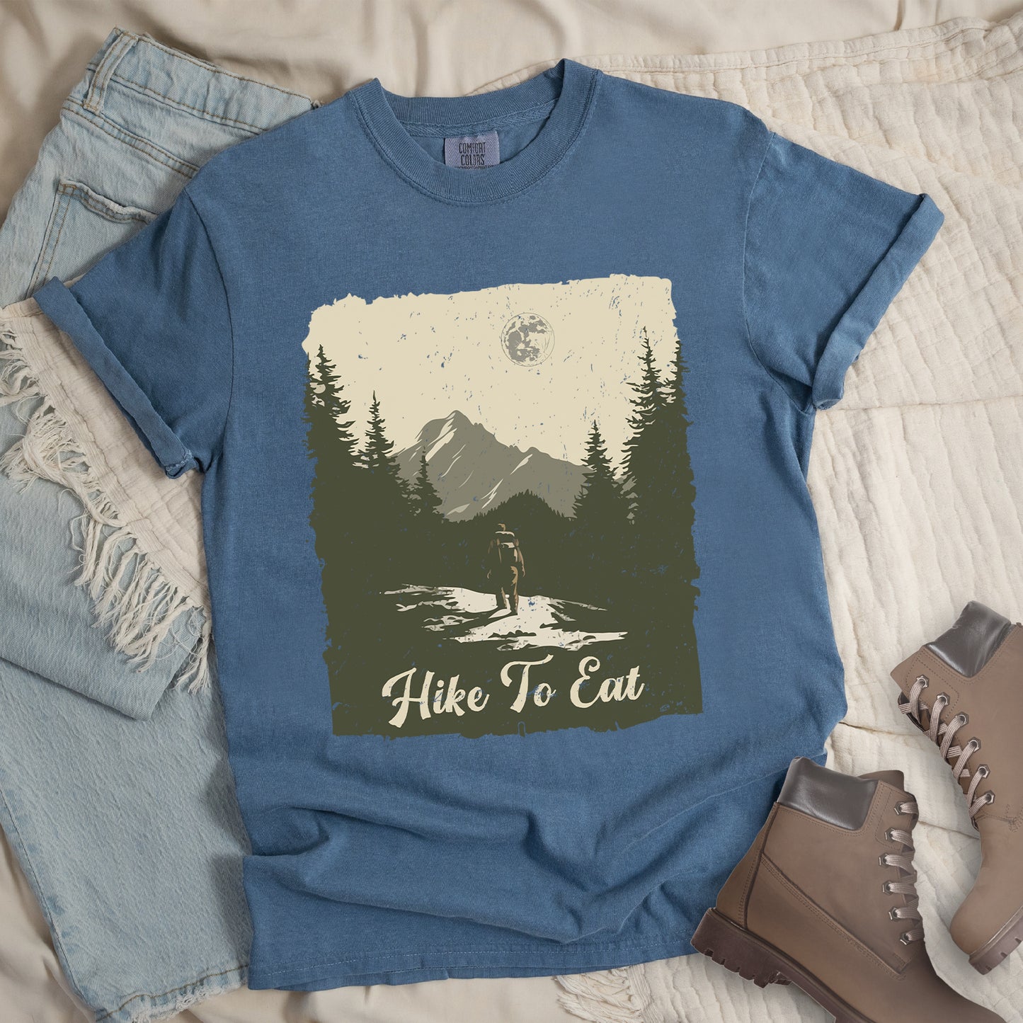 Blue Jean (Denim Blue) t-shirt featuring a vintage-style illustration of a lone hiker on a mountain trail. The rectangular design shows pine trees, mountain silhouettes, and a full moon against a cream-colored sky. A solitary hiker with backpack stands on the path facing the mountains. "Hike To Eat" appears in cream script at the bottom of the illustration. The artwork uses muted green, cream, and gray tones in a distressed, rustic style.