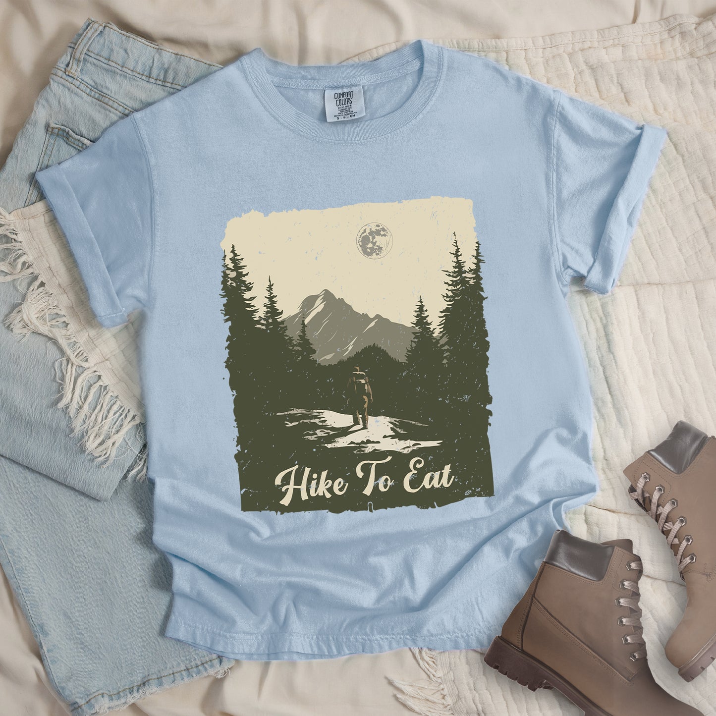 Chambray (Light Blue) t-shirt featuring a vintage-style illustration of a lone hiker on a mountain trail. The rectangular design shows pine trees, mountain silhouettes, and a full moon against a cream-colored sky. A solitary hiker with backpack stands on the path facing the mountains. "Hike To Eat" appears in cream script at the bottom of the illustration. The artwork uses muted green, cream, and gray tones in a distressed, rustic style.