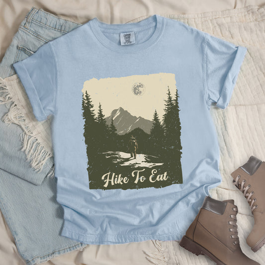 Chambray (Light Blue) t-shirt featuring a vintage-style illustration of a lone hiker on a mountain trail. The rectangular design shows pine trees, mountain silhouettes, and a full moon against a cream-colored sky. A solitary hiker with backpack stands on the path facing the mountains. "Hike To Eat" appears in cream script at the bottom of the illustration. The artwork uses muted green, cream, and gray tones in a distressed, rustic style.