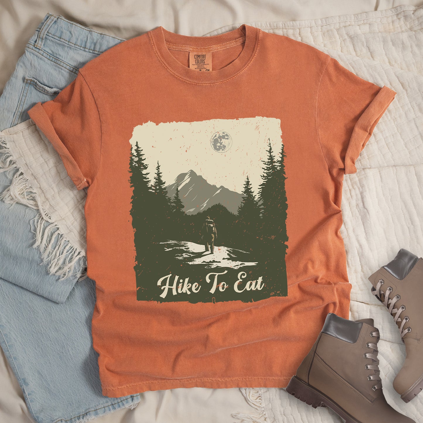 Yam (Brown) t-shirt featuring a vintage-style illustration of a lone hiker on a mountain trail. The rectangular design shows pine trees, mountain silhouettes, and a full moon against a cream-colored sky. A solitary hiker with backpack stands on the path facing the mountains. "Hike To Eat" appears in cream script at the bottom of the illustration. The artwork uses muted green, cream, and gray tones in a distressed, rustic style. 