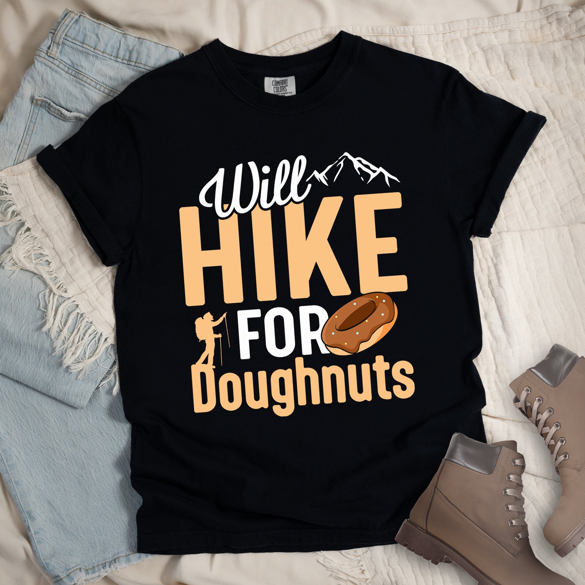 Black t-shirt with "Will HIKE FOR Doughnuts" graphic design. White script "Will" above tan "HIKE" in bold letters, with small mountain peaks above. "FOR" appears in white, followed by "Doughnuts" in tan, with an illustrated chocolate donut replacing the "O" in "FOR". Features a small hiker silhouette.  The shirt features a classic crew neck and casual rolled sleeves, styled alongside light wash denim and brown hiking boots. 