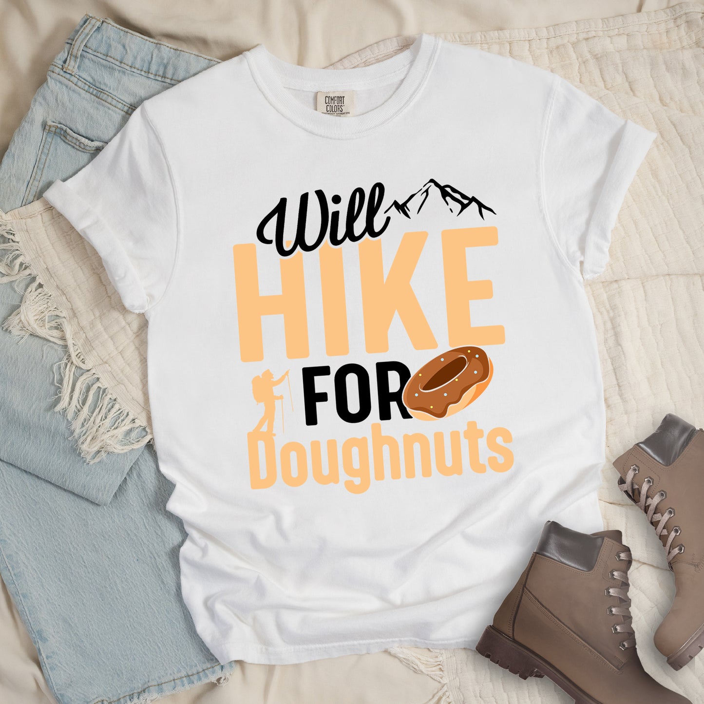 White t-shirt with "Will HIKE FOR Doughnuts" graphic design. Black script "Will" above tan "HIKE" in bold letters, with small mountain peaks above. "FOR" appears in black, followed by "Doughnuts" in tan, with an illustrated chocolate donut replacing the "O" in "FOR". Features a small hiker silhouette. The shirt features a classic crew neck and casual rolled sleeves, styled alongside light wash denim and brown hiking boots. 