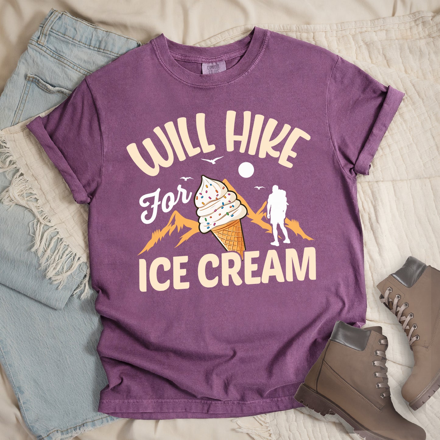 Berry (Dark Violet) t-shirt with cream-colored text reading "WILL HIKE For ICE CREAM". "WILL HIKE" appears in an arc at the top, "For" in white script in the middle, and "ICE CREAM" in cream letters at the bottom. The design features orange mountain silhouettes, a white hiker figure, flying birds, a sun/moon, and an illustration of an ice cream cone with sprinkles. The design combines hiking motivation with dessert rewards.