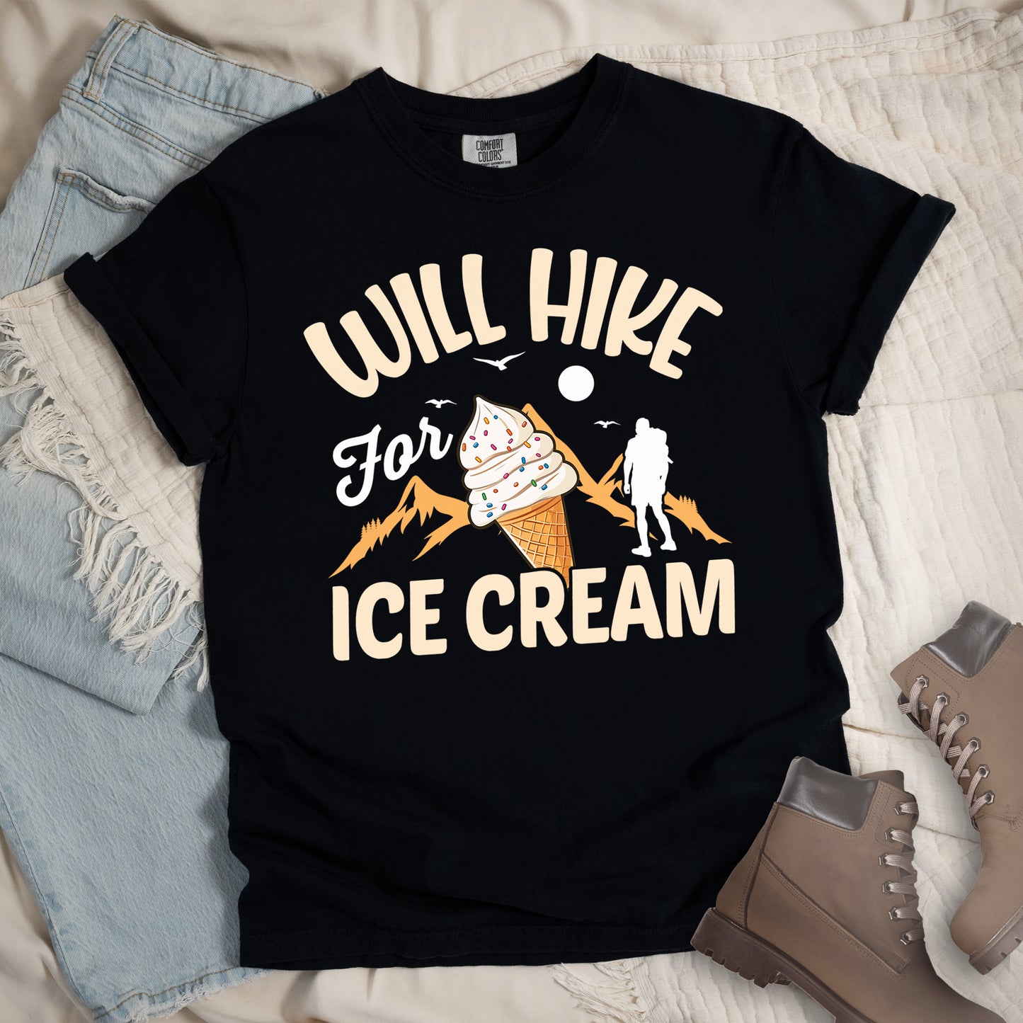 Black t-shirt with cream-colored text reading "WILL HIKE For ICE CREAM". "WILL HIKE" appears in an arc at the top, "For" in white script in the middle, and "ICE CREAM" in cream letters at the bottom. The design features orange mountain silhouettes, a white hiker figure, flying birds, a sun/moon, and an illustration of an ice cream cone with sprinkles. The design combines hiking motivation with dessert rewards.