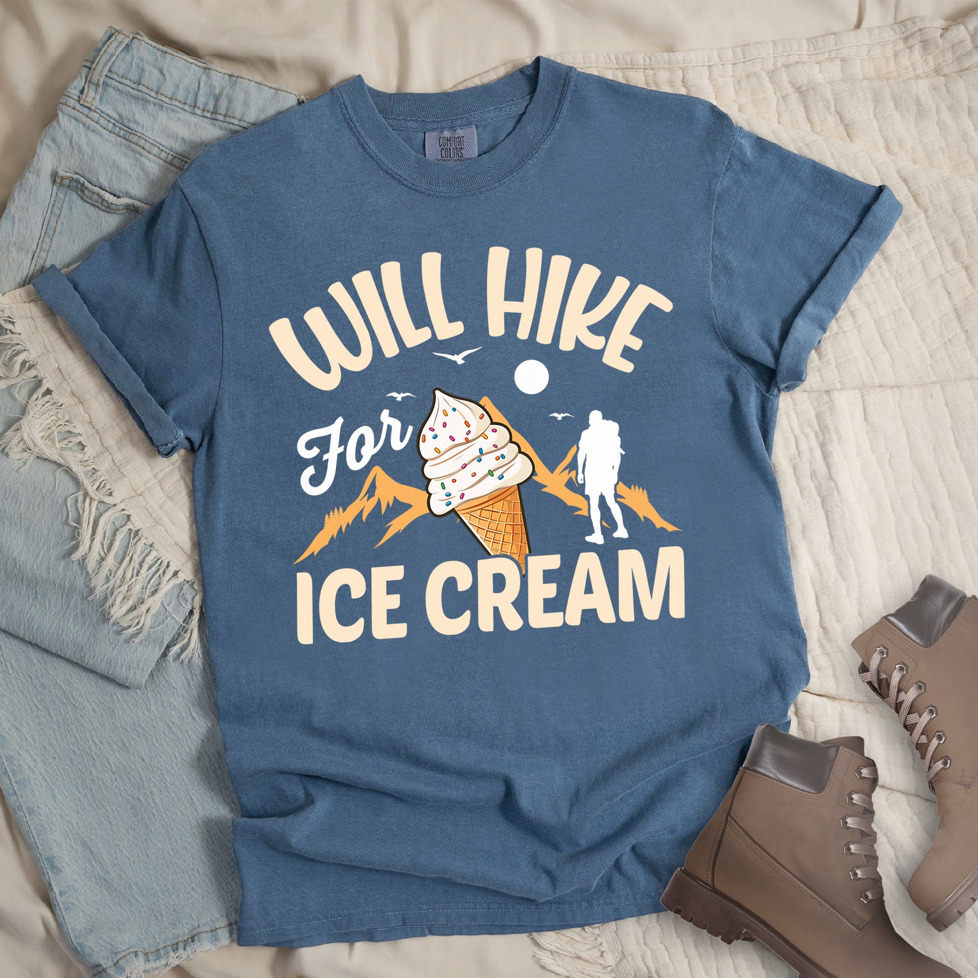 Blue Jean (Denim Blue) t-shirt with cream-colored text reading "WILL HIKE For ICE CREAM". "WILL HIKE" appears in an arc at the top, "For" in white script in the middle, and "ICE CREAM" in cream letters at the bottom. The design features orange mountain silhouettes, a white hiker figure, flying birds, a sun/moon, and an illustration of an ice cream cone with sprinkles. The design combines hiking motivation with dessert rewards.