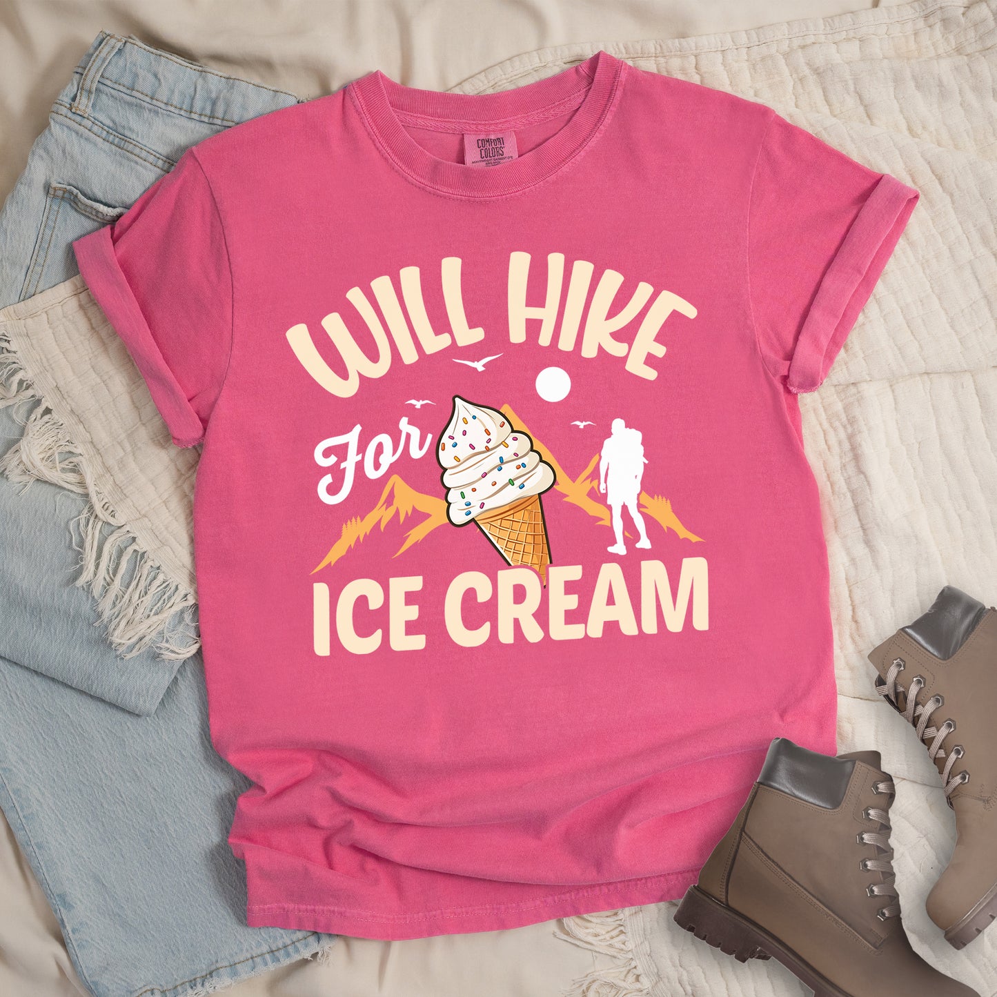 Crunchberry (Bright Pink) t-shirt with cream-colored text reading "WILL HIKE For ICE CREAM". "WILL HIKE" appears in an arc at the top, "For" in white script in the middle, and "ICE CREAM" in cream letters at the bottom. The design features orange mountain silhouettes, a white hiker figure, flying birds, a sun/moon, and an illustration of an ice cream cone with sprinkles. The design combines hiking motivation with dessert rewards.