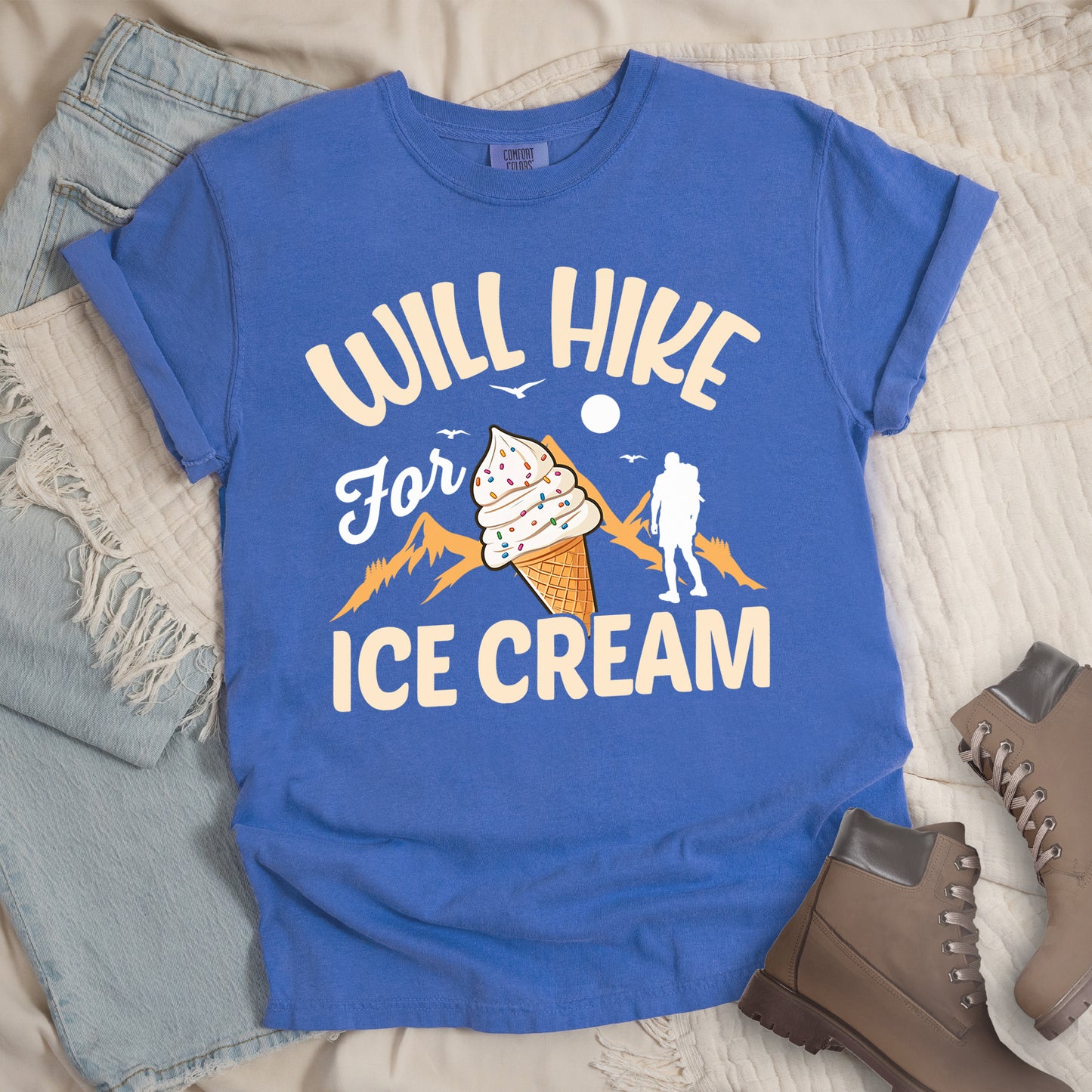 Flo Blue (Bright Blue) t-shirt with cream-colored text reading "WILL HIKE For ICE CREAM". "WILL HIKE" appears in an arc at the top, "For" in white script in the middle, and "ICE CREAM" in cream letters at the bottom. The design features orange mountain silhouettes, a white hiker figure, flying birds, a sun/moon, and an illustration of an ice cream cone with sprinkles. The design combines hiking motivation with dessert rewards.