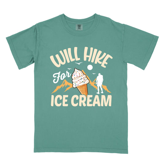 Light Green t-shirt with cream-colored text reading "WILL HIKE For ICE CREAM". "WILL HIKE" appears in an arc at the top, "For" in white script in the middle, and "ICE CREAM" in cream letters at the bottom. The design features orange mountain silhouettes, a white hiker figure, flying birds, a sun/moon, and an illustration of an ice cream cone with sprinkles. The design combines hiking motivation with dessert rewards.
