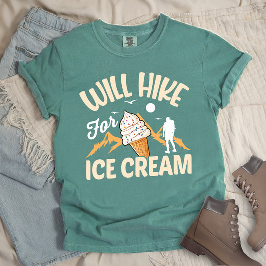 Light Green t-shirt with cream-colored text reading "WILL HIKE For ICE CREAM". "WILL HIKE" appears in an arc at the top, "For" in white script in the middle, and "ICE CREAM" in cream letters at the bottom. The design features orange mountain silhouettes, a white hiker figure, flying birds, a sun/moon, and an illustration of an ice cream cone with sprinkles. The design combines hiking motivation with dessert rewards.