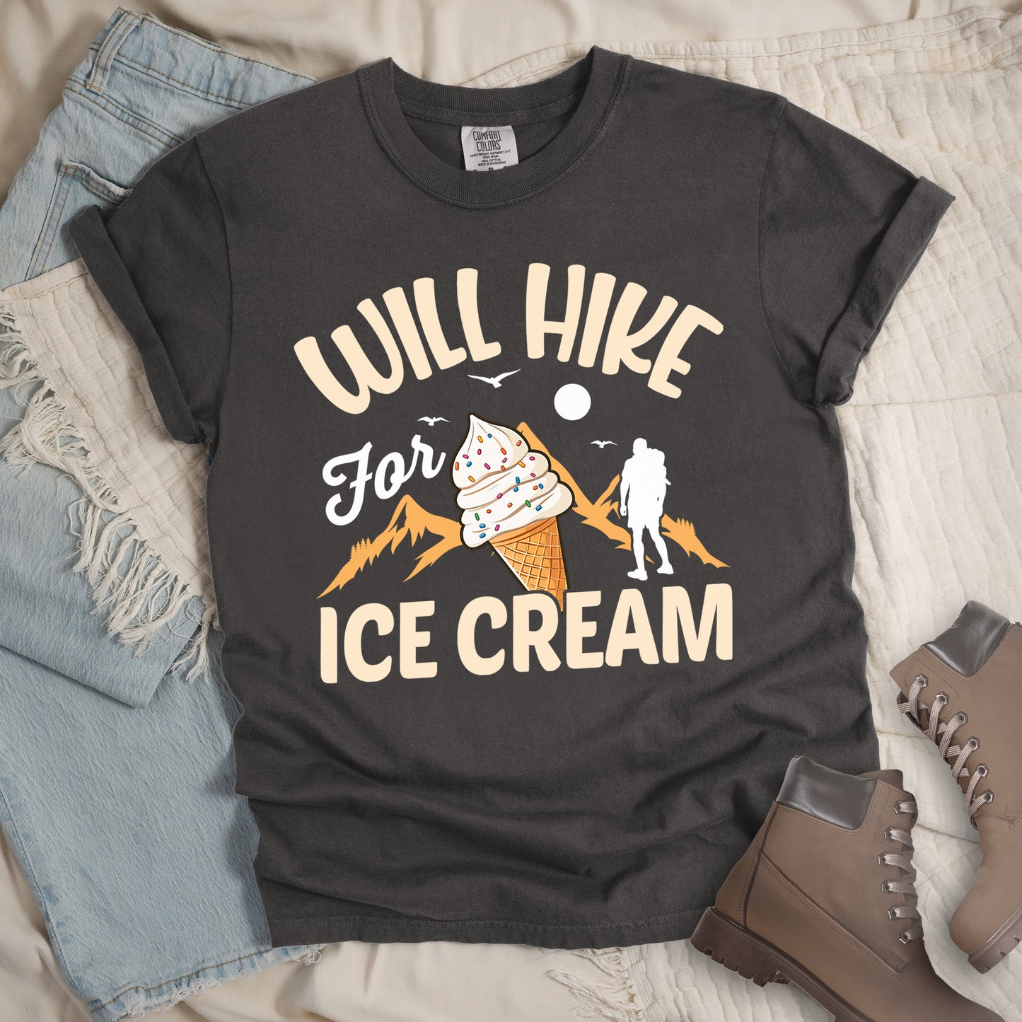 Pepper (Dark Gray) t-shirt with cream-colored text reading "WILL HIKE For ICE CREAM". "WILL HIKE" appears in an arc at the top, "For" in white script in the middle, and "ICE CREAM" in cream letters at the bottom. The design features orange mountain silhouettes, a white hiker figure, flying birds, a sun/moon, and an illustration of an ice cream cone with sprinkles. The design combines hiking motivation with dessert rewards.