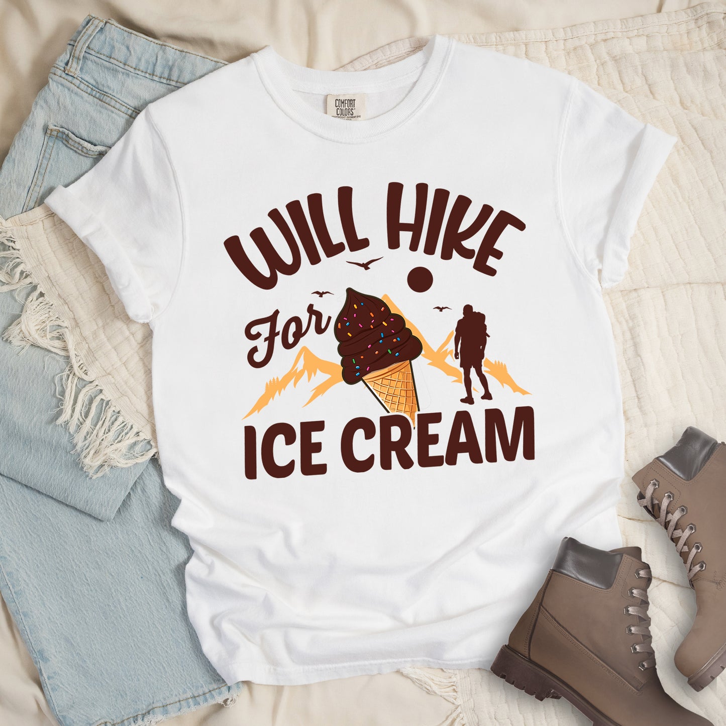 White t-shirt with deep brown-colored text reading "WILL HIKE For ICE CREAM". "WILL HIKE" appears in an arc at the top, "For" in deep brown script in the middle, and "ICE CREAM" in deep brown letters at the bottom. The design features orange mountain silhouettes, a deep brown hiker figure, flying birds, a sun/moon, and an illustration of an ice cream cone with sprinkles. The design combines hiking motivation with dessert rewards.