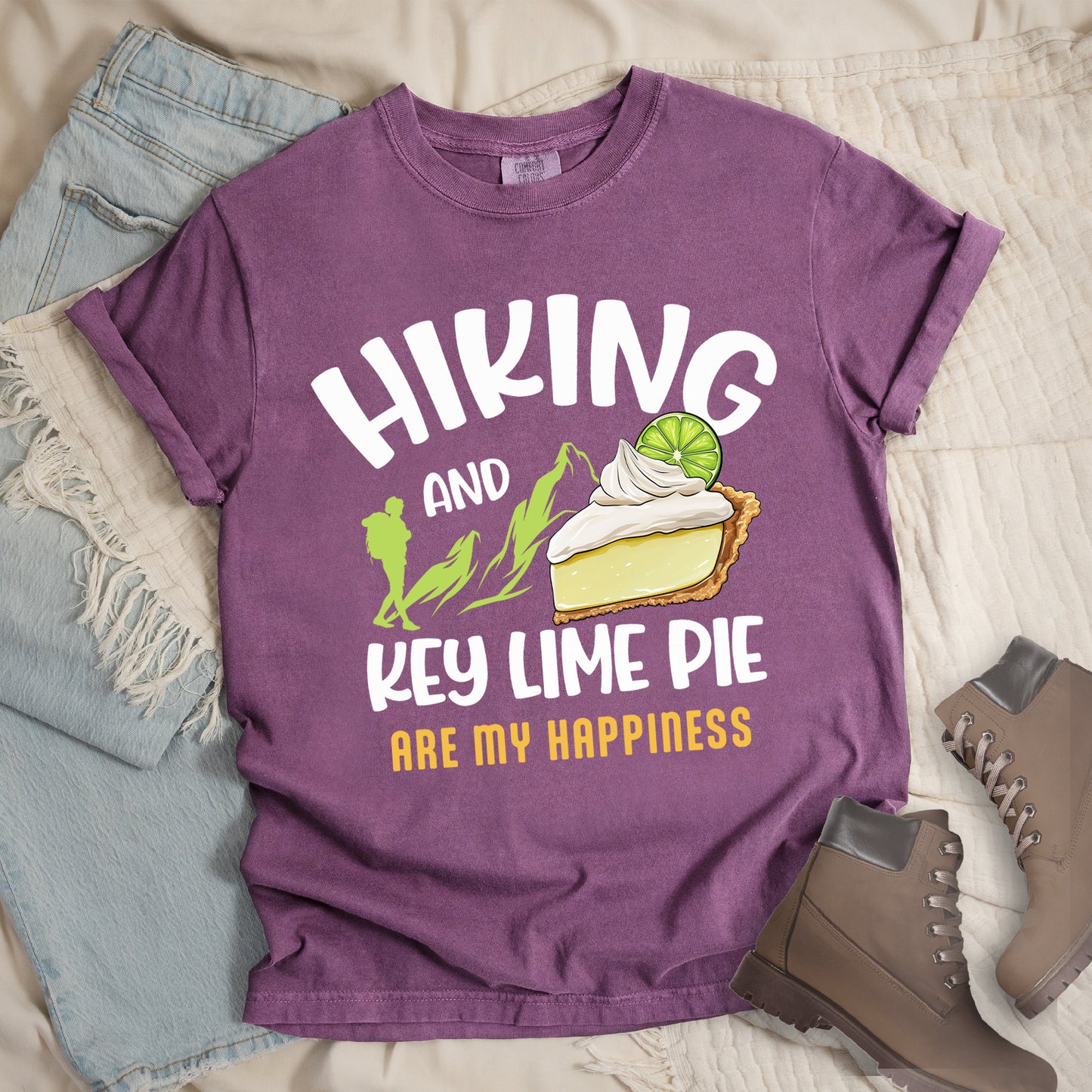 Berry (Dark Violet) t-shirt with text "HIKING AND KEY LIME PIE ARE MY HAPPINESS". "HIKING" appears in white arched letters at the top, "AND" in small white text, "KEY LIME PIE" in large white text in the middle, and "ARE MY HAPPINESS" in gold/yellow at the bottom. The design includes green mountain silhouettes with a hiker silhouette and an illustration of a slice of key lime pie with whipped cream and lime garnish in between "AND" and "KEY LIME PIE." 