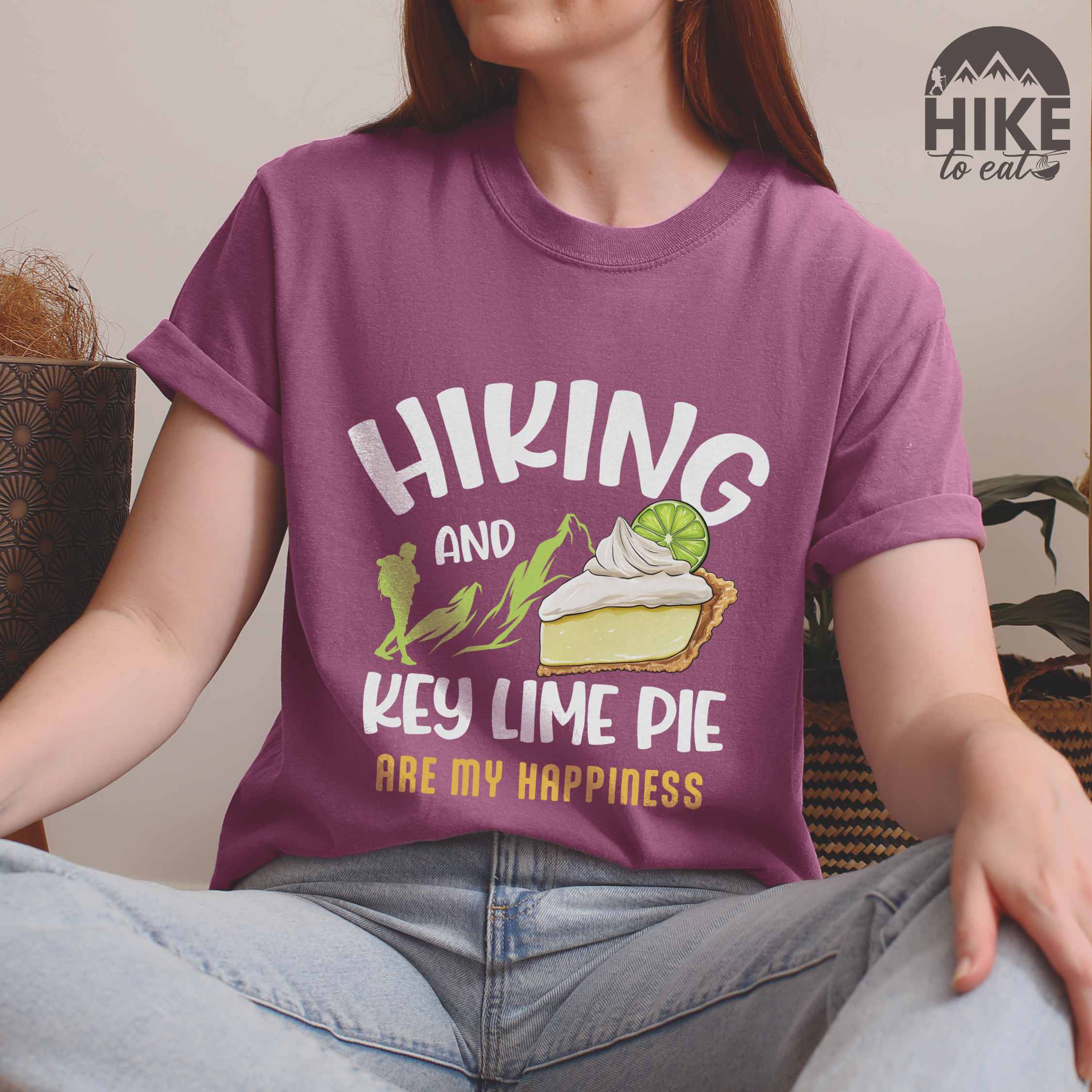 Woman wearing berry "Hiking and key lime pie are my happiness" colored vintage tee with bold typography, mountains, birds, illustrated slice of key lime pie, and backpacker for pie lovers.