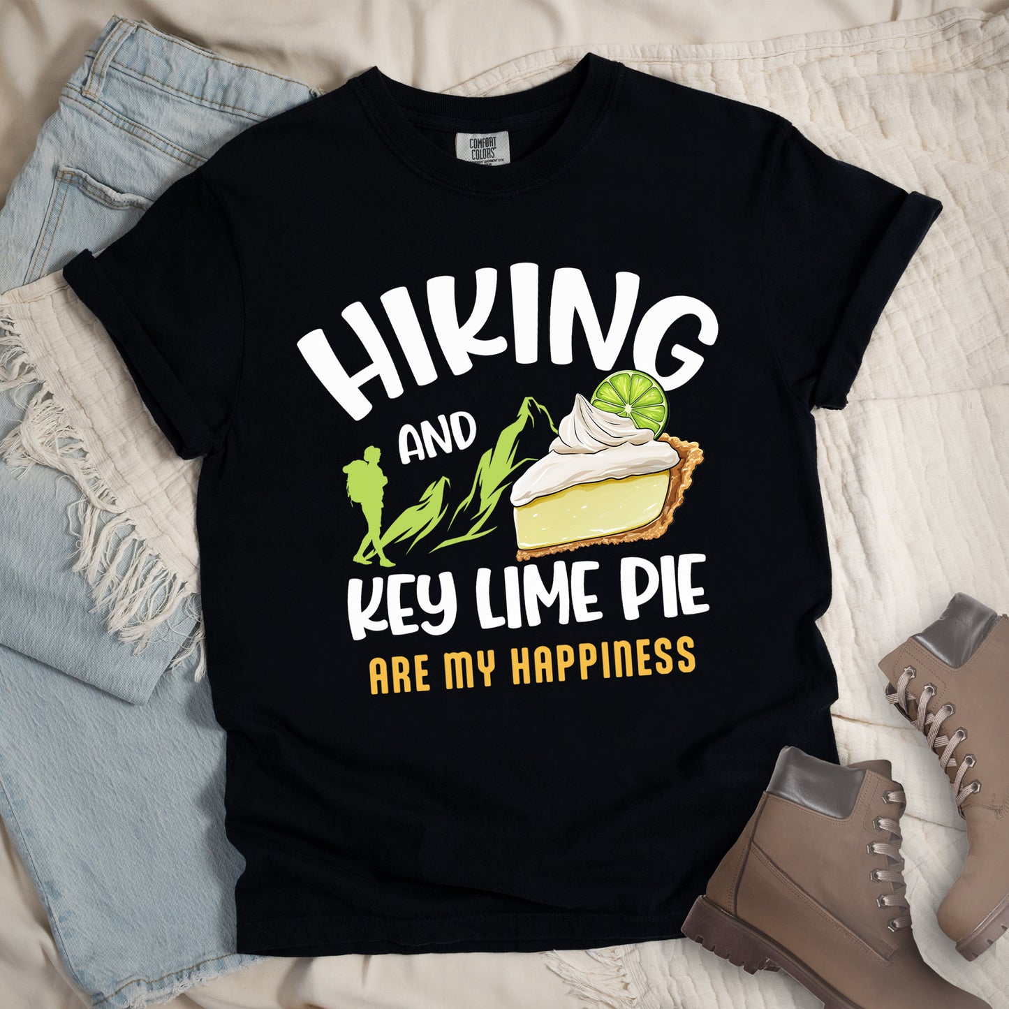Black t-shirt with text "HIKING AND KEY LIME PIE ARE MY HAPPINESS". "HIKING" appears in white arched letters at the top, "AND" in small white text, "KEY LIME PIE" in large white text in the middle, and "ARE MY HAPPINESS" in gold/yellow at the bottom. The design includes green mountain silhouettes with a hiker silhouette and an illustration of a slice of key lime pie with whipped cream and lime garnish in between "AND" and "KEY LIME PIE." 