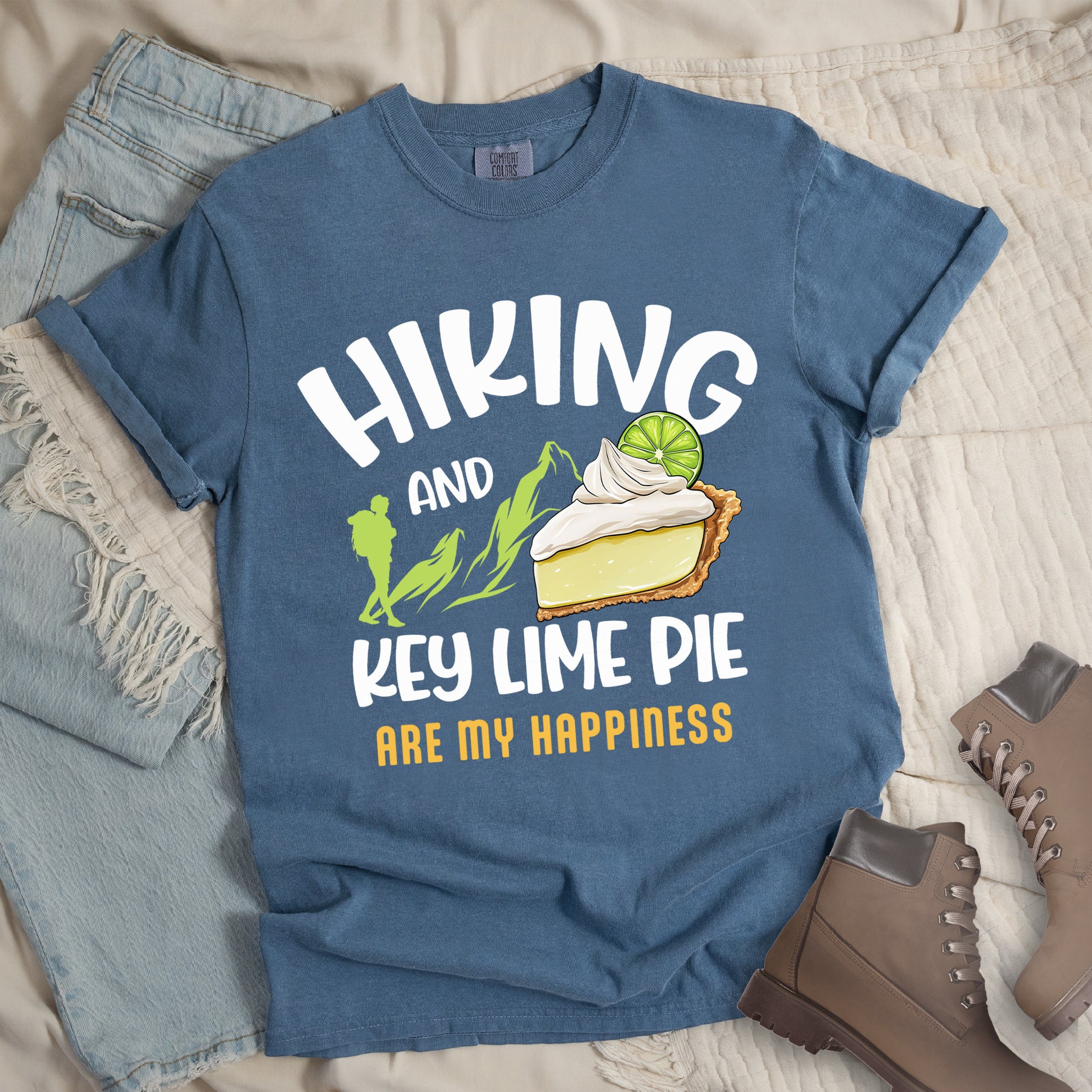 Blue Jean (Denim Blue) t-shirt with text "HIKING AND KEY LIME PIE ARE MY HAPPINESS". "HIKING" appears in white arched letters at the top, "AND" in small white text, "KEY LIME PIE" in large white text in the middle, and "ARE MY HAPPINESS" in gold/yellow at the bottom. The design includes green mountain silhouettes with a hiker silhouette and an illustration of a slice of key lime pie with whipped cream and lime garnish in between "AND" and "KEY LIME PIE." 