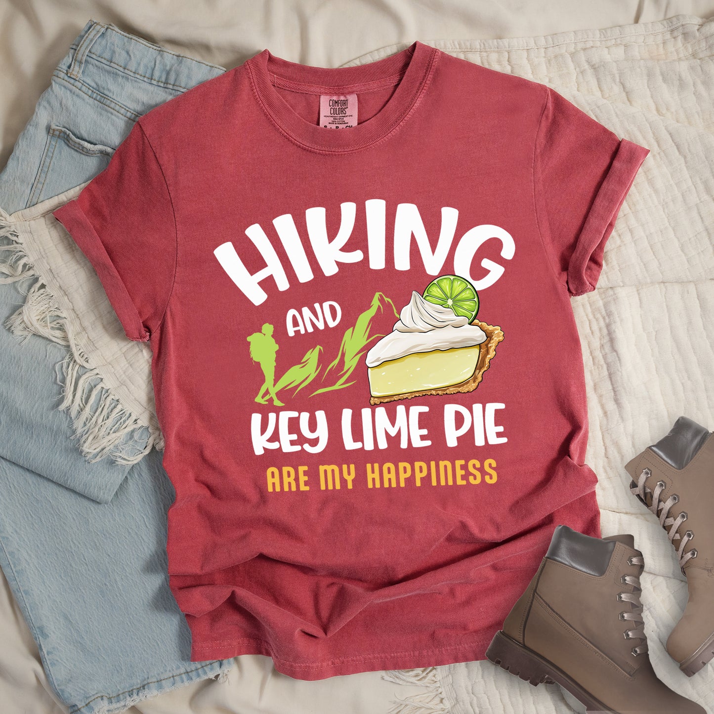 Crimson (Dark Pink) t-shirt with text "HIKING AND KEY LIME PIE ARE MY HAPPINESS". "HIKING" appears in white arched letters at the top, "AND" in small white text, "KEY LIME PIE" in large white text in the middle, and "ARE MY HAPPINESS" in gold/yellow at the bottom. The design includes green mountain silhouettes with a hiker silhouette and an illustration of a slice of key lime pie with whipped cream and lime garnish in between "AND" and "KEY LIME PIE." 