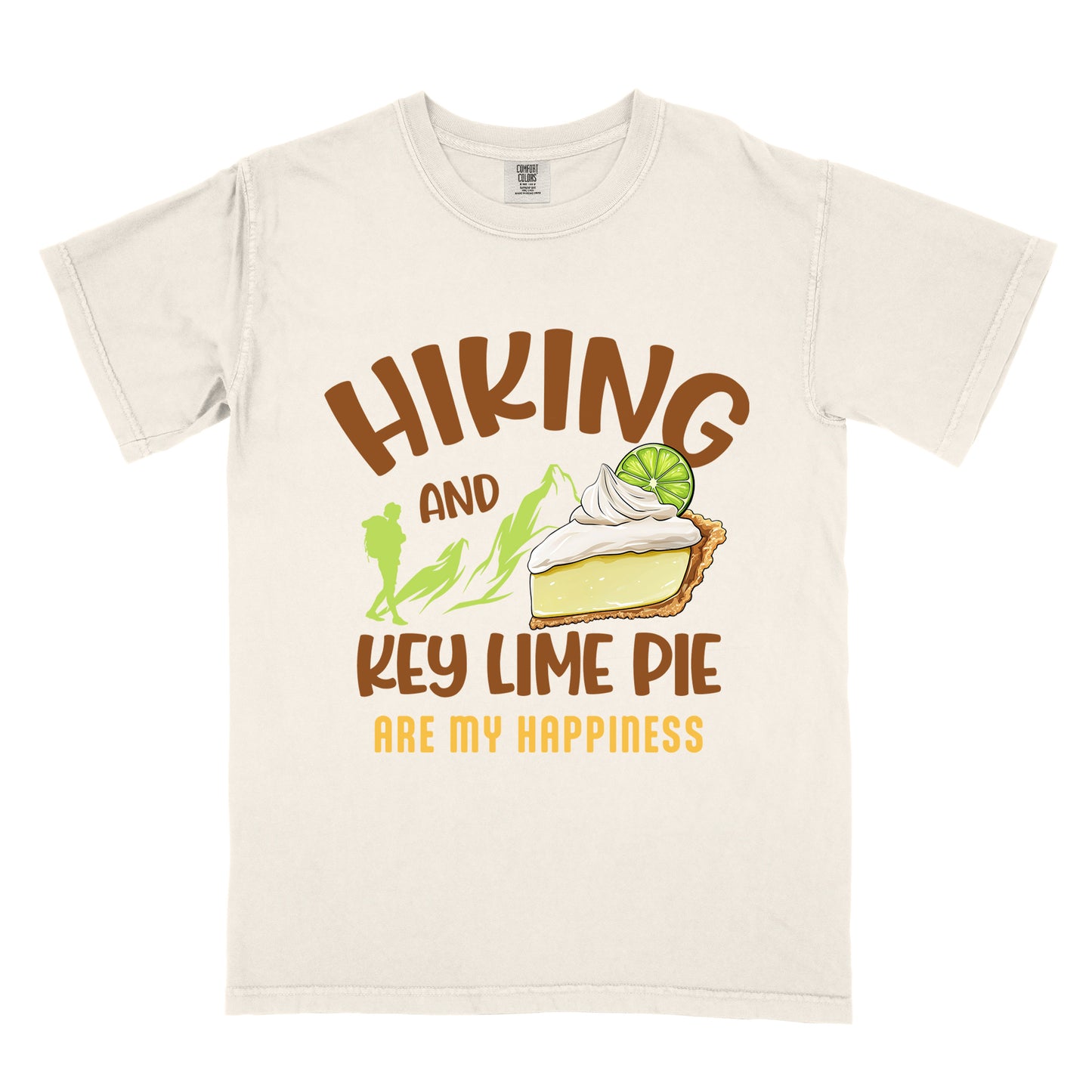 Ivory (Cream White) t-shirt with text "HIKING AND KEY LIME PIE ARE MY HAPPINESS". "HIKING" appears in brown arched letters at the top, "AND" in small brown text, "KEY LIME PIE" in large brown text in the middle, and "ARE MY HAPPINESS" in gold/yellow at the bottom. The design includes green mountain silhouettes with a hiker silhouette and an illustration of a slice of key lime pie with whipped cream and lime garnish in between "AND" and "KEY LIME PIE." 