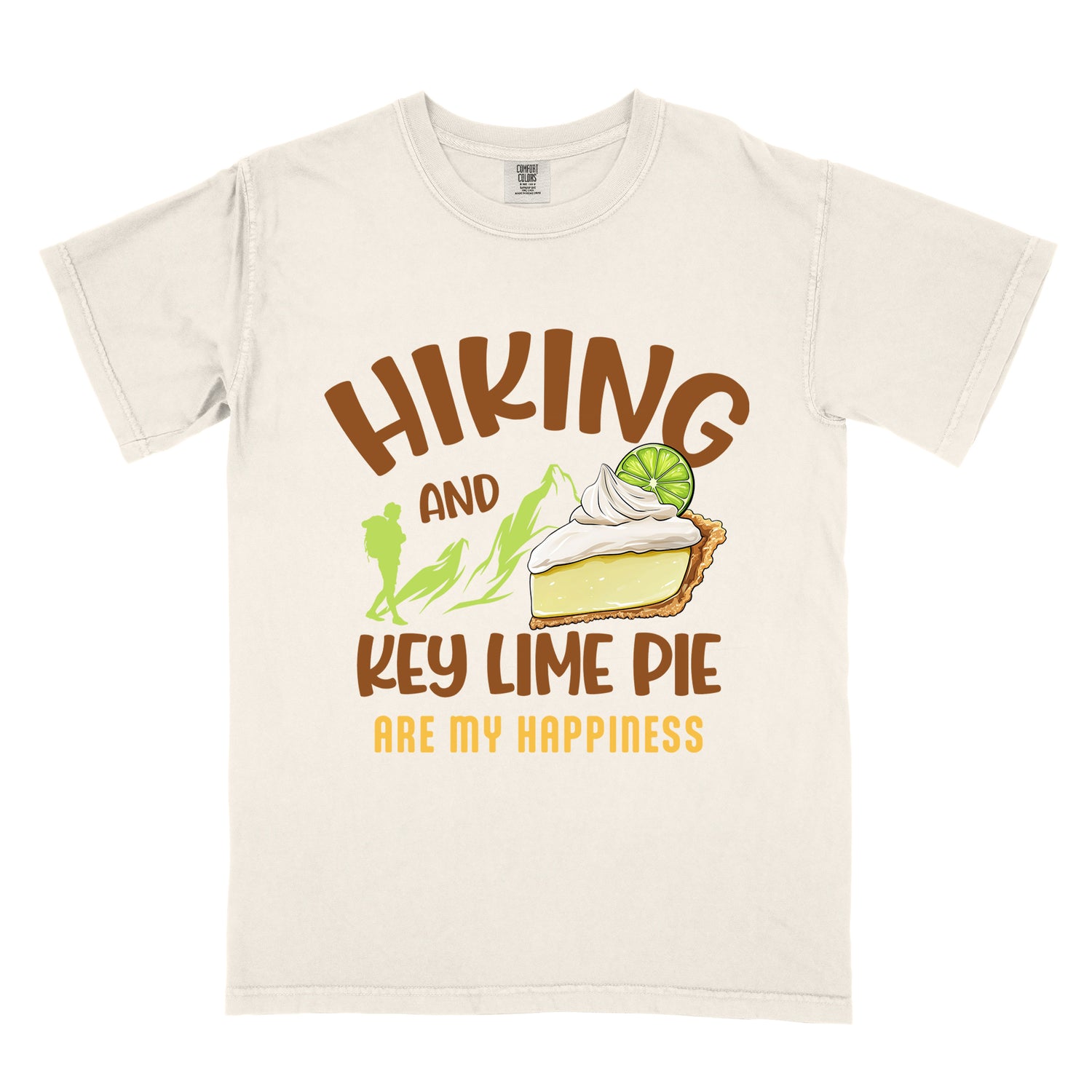 Ivory (Cream White) t-shirt with text "HIKING AND KEY LIME PIE ARE MY HAPPINESS". "HIKING" appears in brown arched letters at the top, "AND" in small brown text, "KEY LIME PIE" in large brown text in the middle, and "ARE MY HAPPINESS" in gold/yellow at the bottom. The design includes green mountain silhouettes with a hiker silhouette and an illustration of a slice of key lime pie with whipped cream and lime garnish in between "AND" and "KEY LIME PIE." 